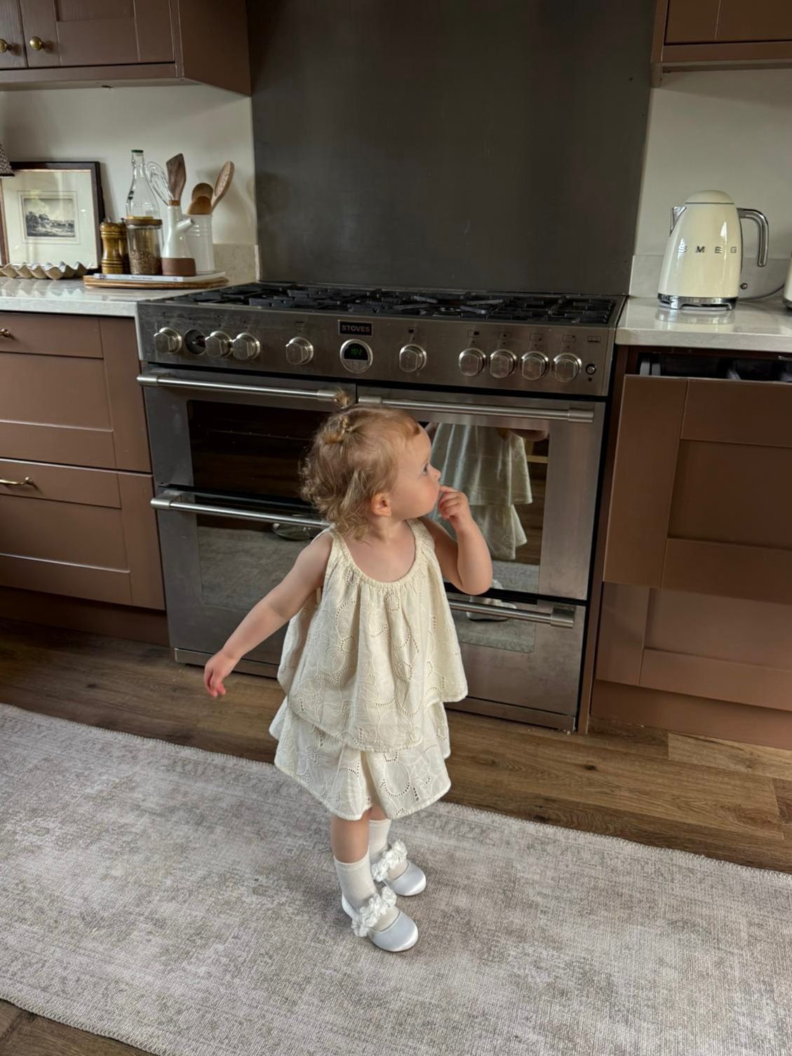 Toddler girl in white dress and socks stands in front of stainless steel double oven range in modern kitchen.