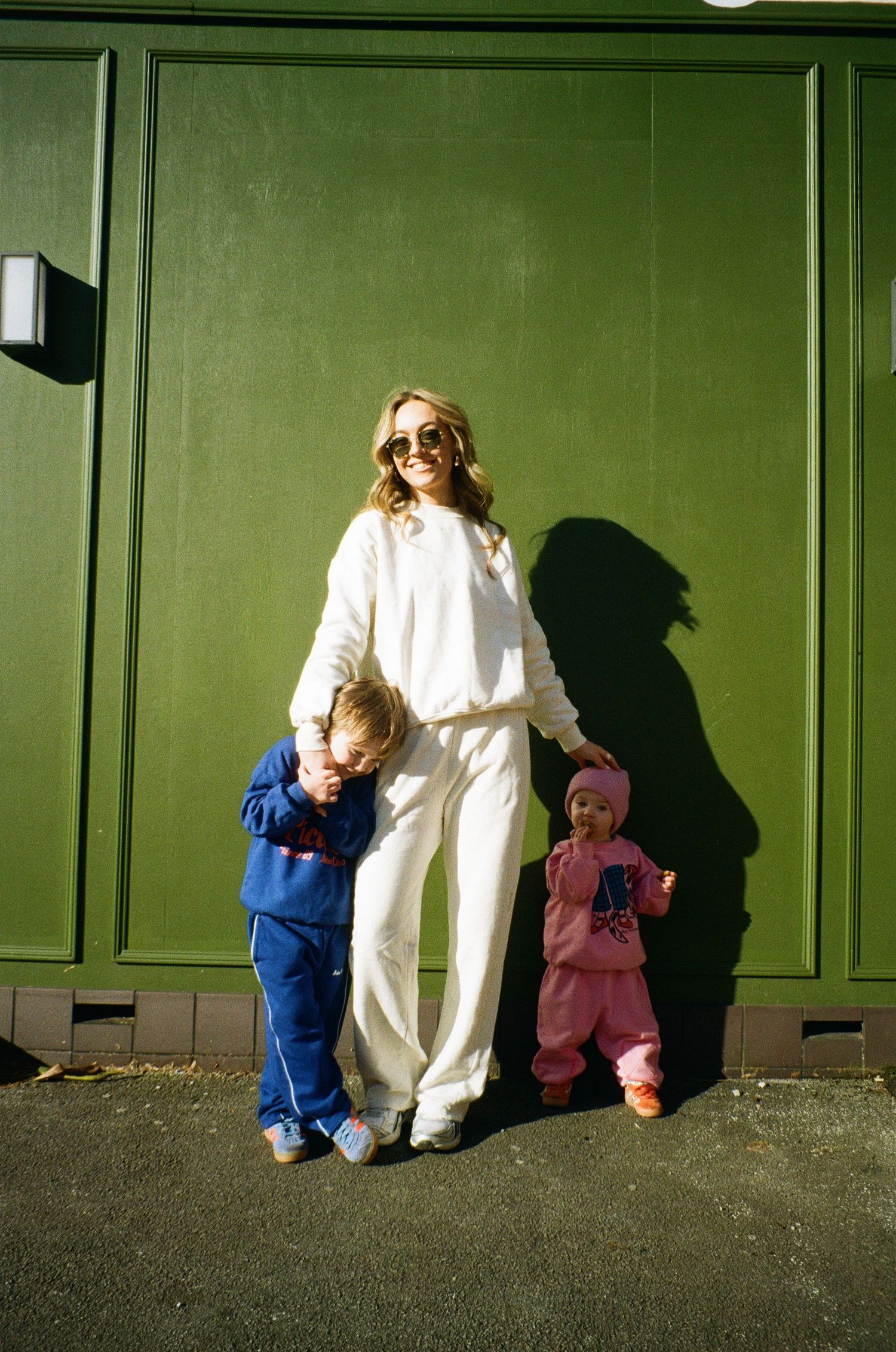 Figure in white outfit stands against green wall with two small children in blue and pink tracksuits.