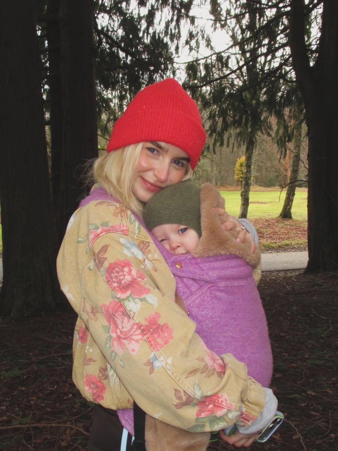 Woman in red beanie carrying a baby in a purple wrap carrier outdoors among trees in cool weather.