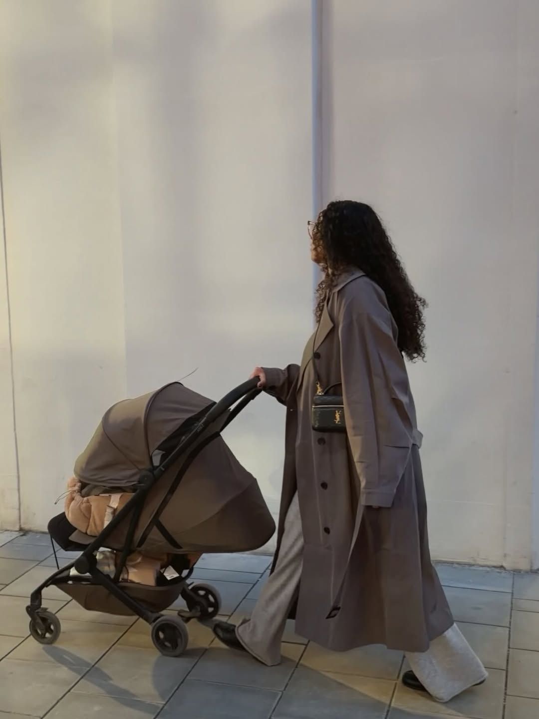 A person in a long beige coat walks past a white wall while pushing a stroller in a minimalist setting.