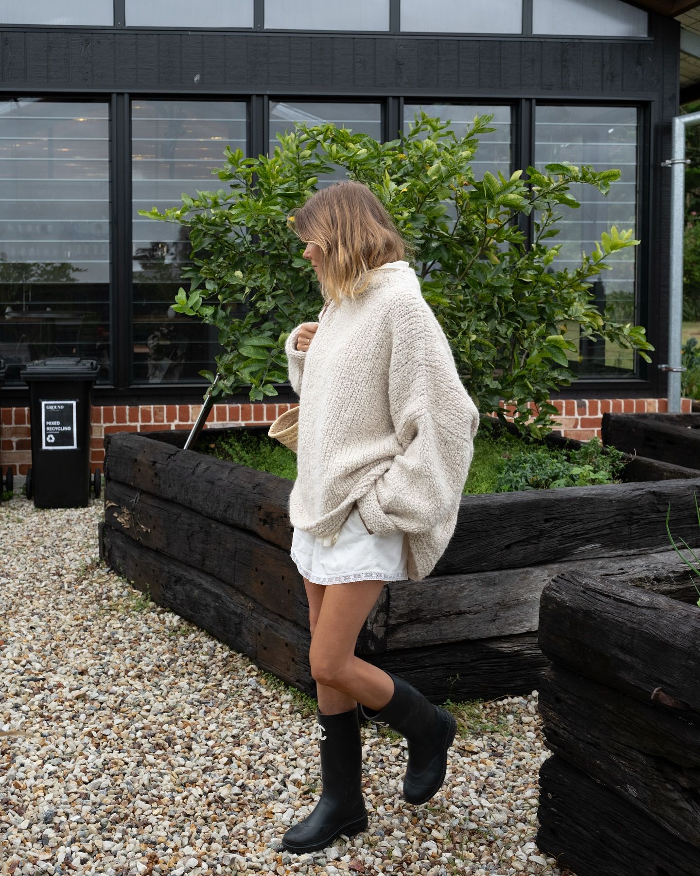 Woman in oversized cream knit sweater, white shorts, and black knee-high boots standing outdoors near wooden planters.