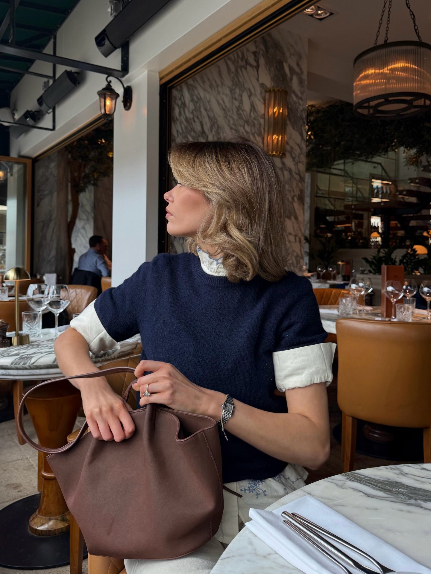 Woman in navy top holding brown leather bag seated at an elegant restaurant table.