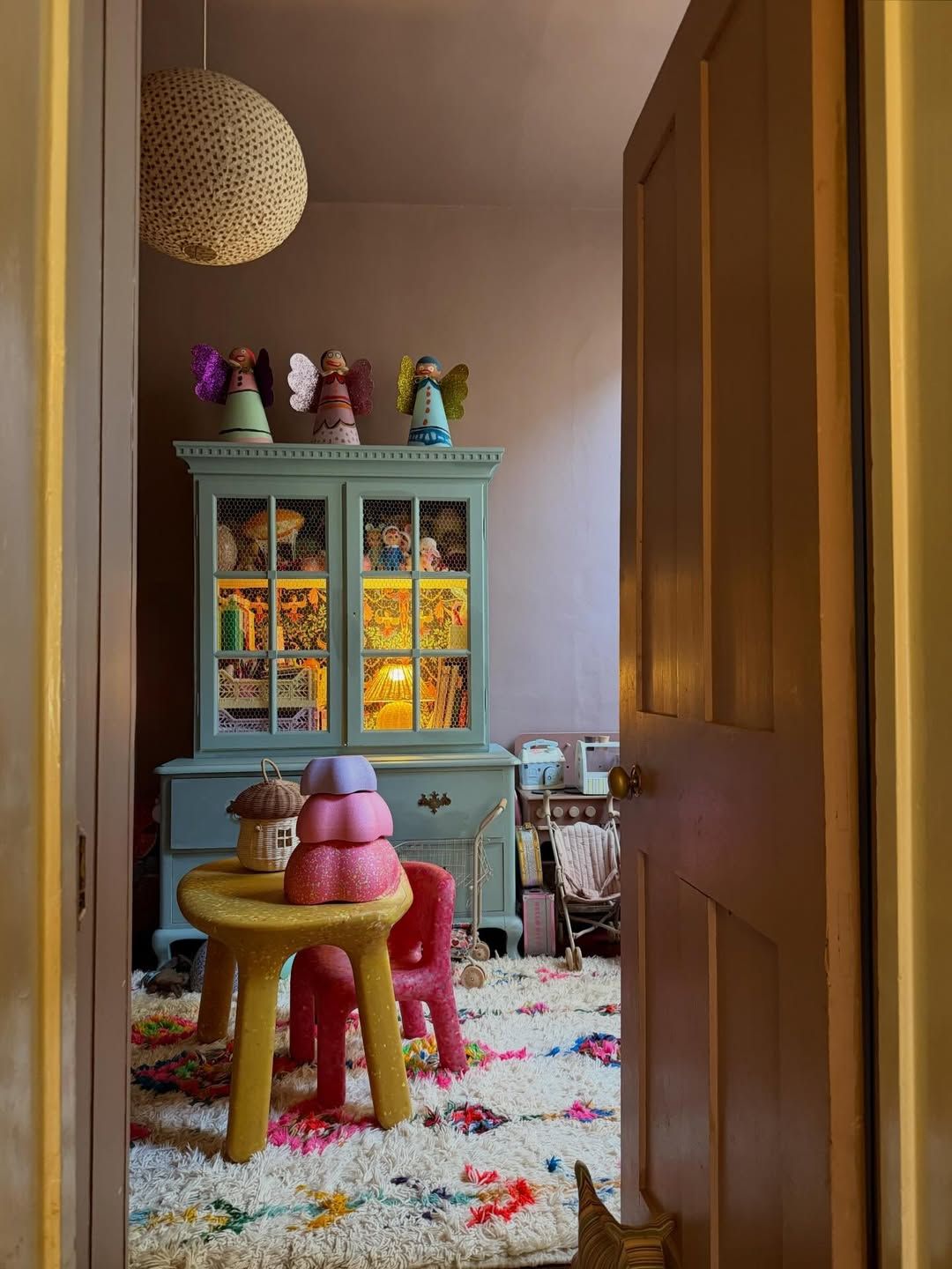 Colorful children's playroom with teal cabinet, yellow stool, pink chair, plush rug, and glowing display case with toys.