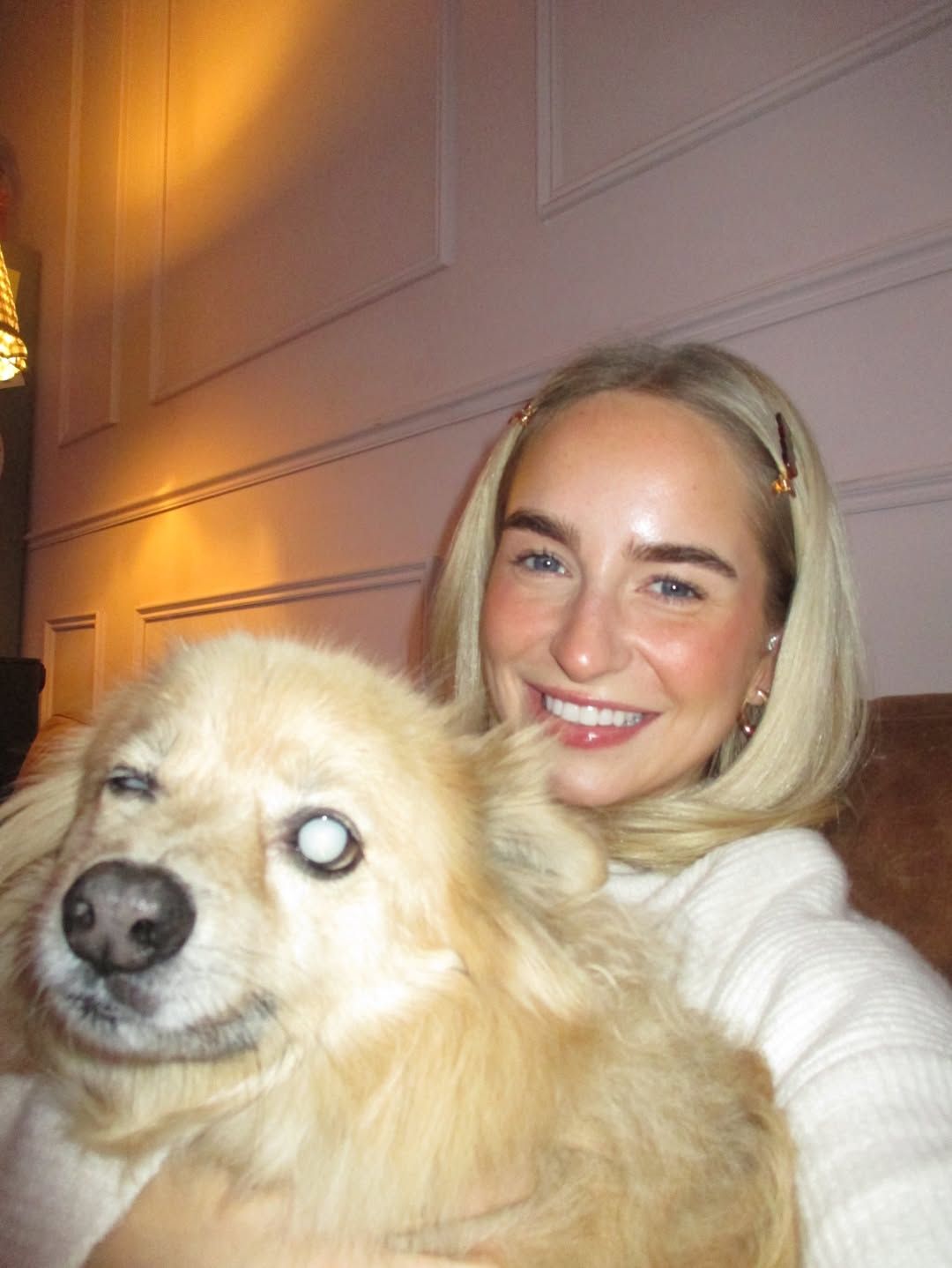 Smiling blonde woman in white sweater taking selfie with fluffy golden dog with cloudy eye on couch.