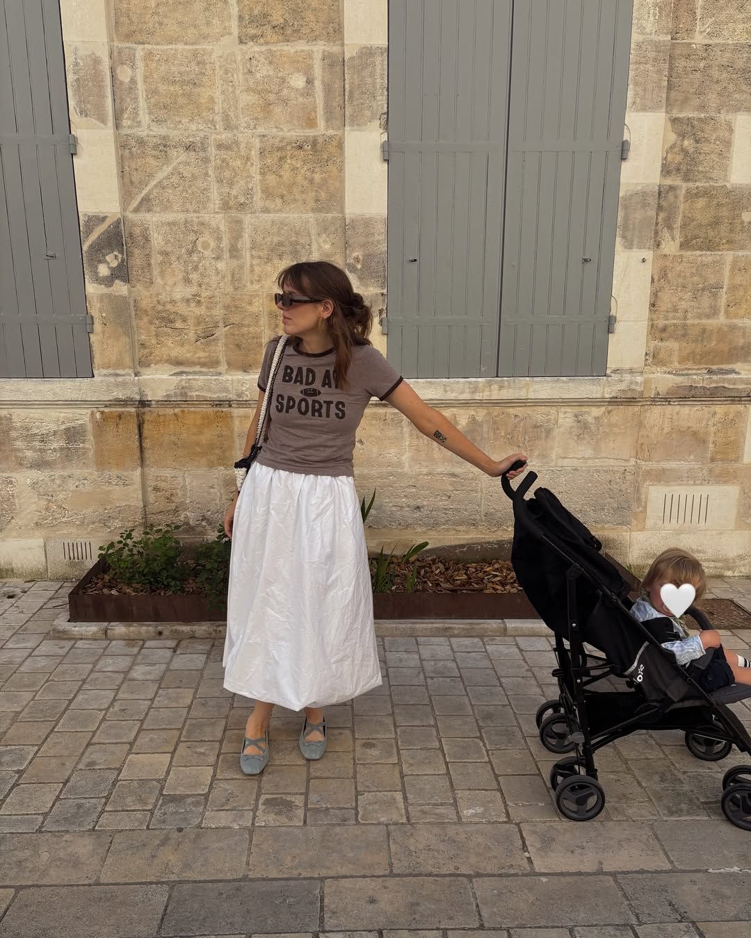 Person in a brown t-shirt and white skirt stands on stone pavement while pushing a stroller near a historic building.