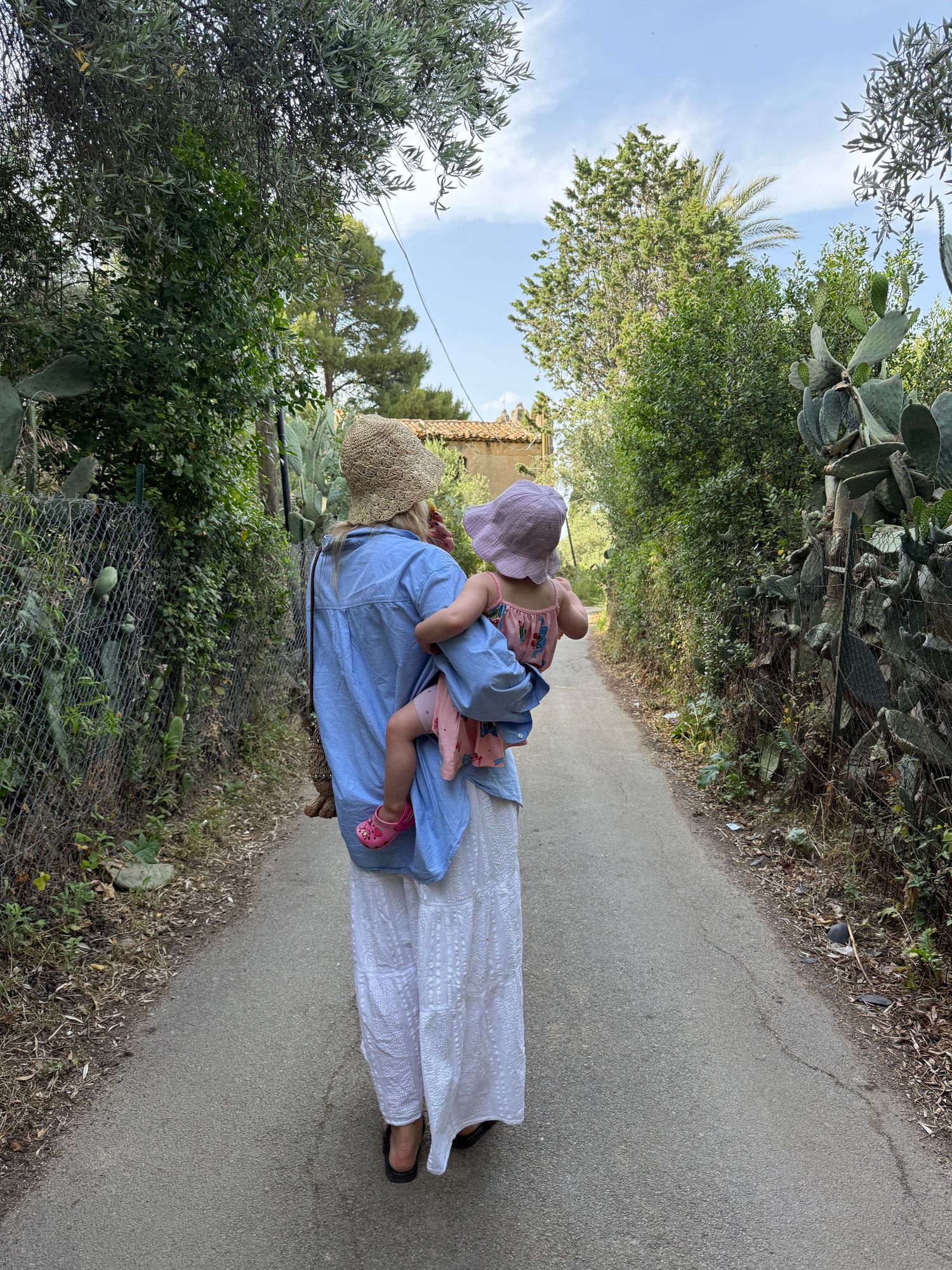 Woman in blue dress and straw hat carries a baby along a narrow Mediterranean path lined with cacti.