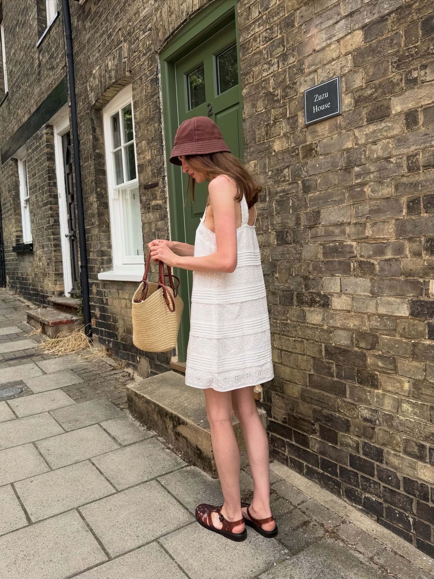 Woman in white sundress, brown bucket hat, and sandals holding a wicker bag outside a brick building.