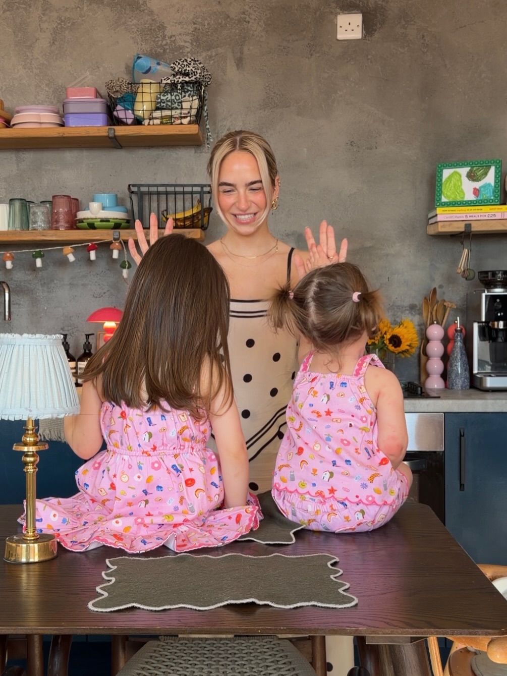 A smiling woman interacts with two young girls in matching pink floral outfits seated on a table in a cozy kitchen.