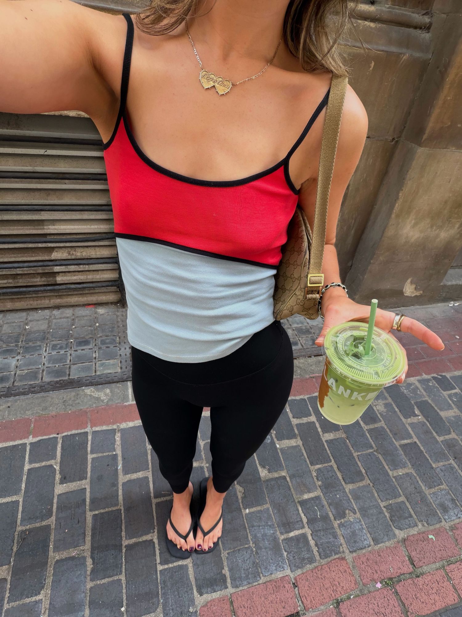 Woman in colorblock tank top and black leggings holds an iced matcha drink on a cobblestone street.