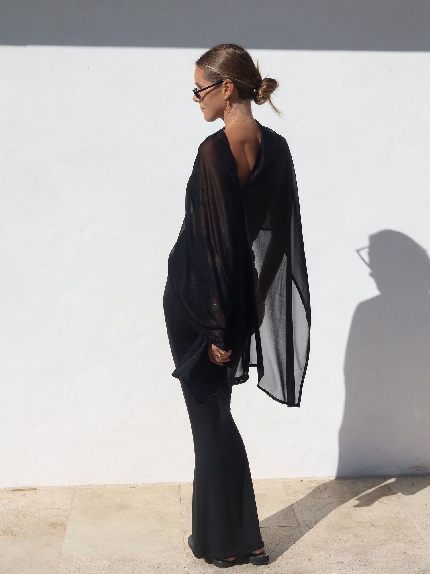 Woman in black sheer oversized top and flared pants poses against white wall in sunlight.