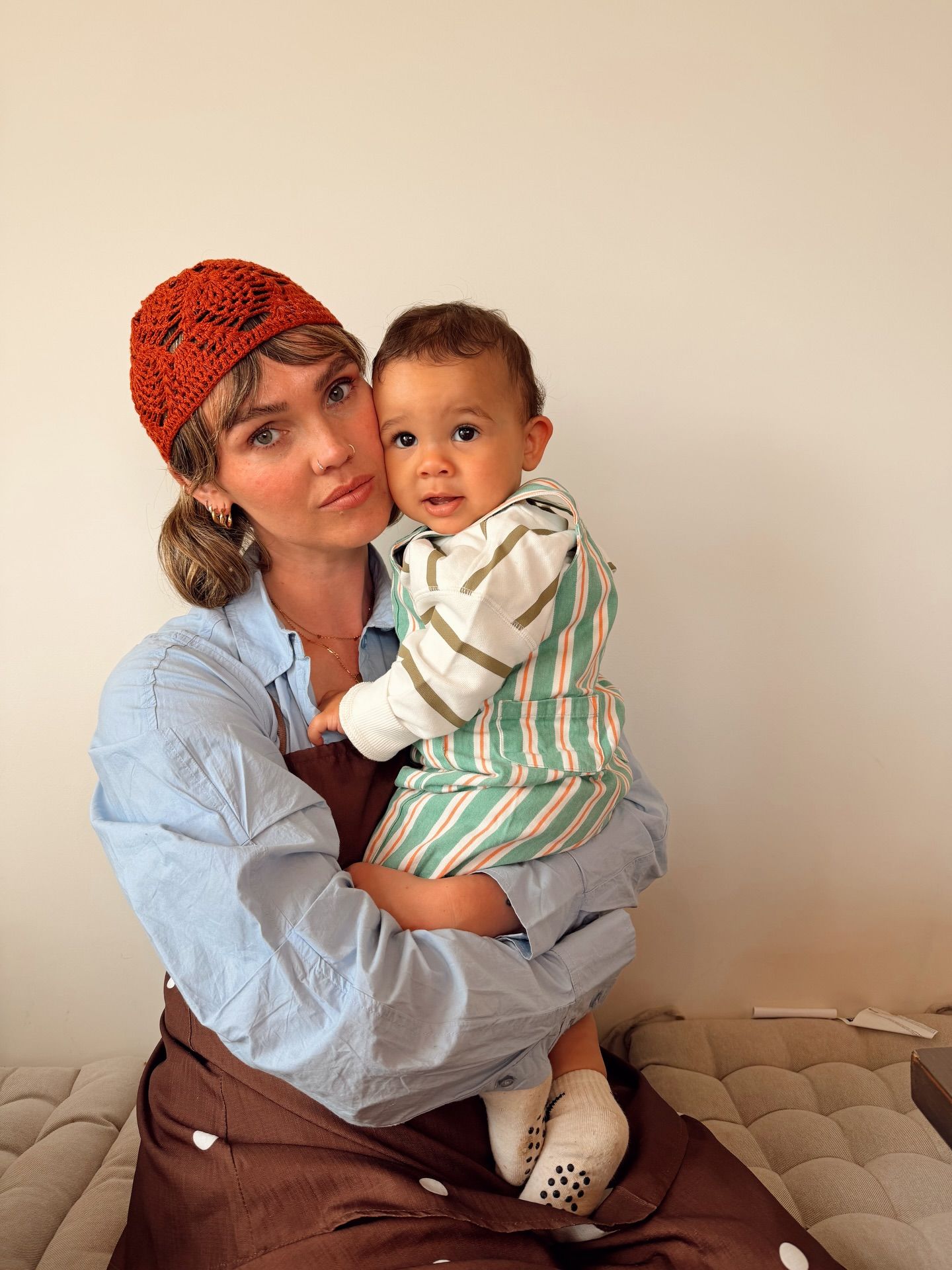 Woman wearing orange crochet hat holds a baby in striped outfit while sitting on a couch.