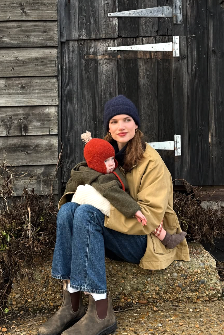 A person in a navy beanie and tan jacket sits against a rustic wooden barn wall in autumn.