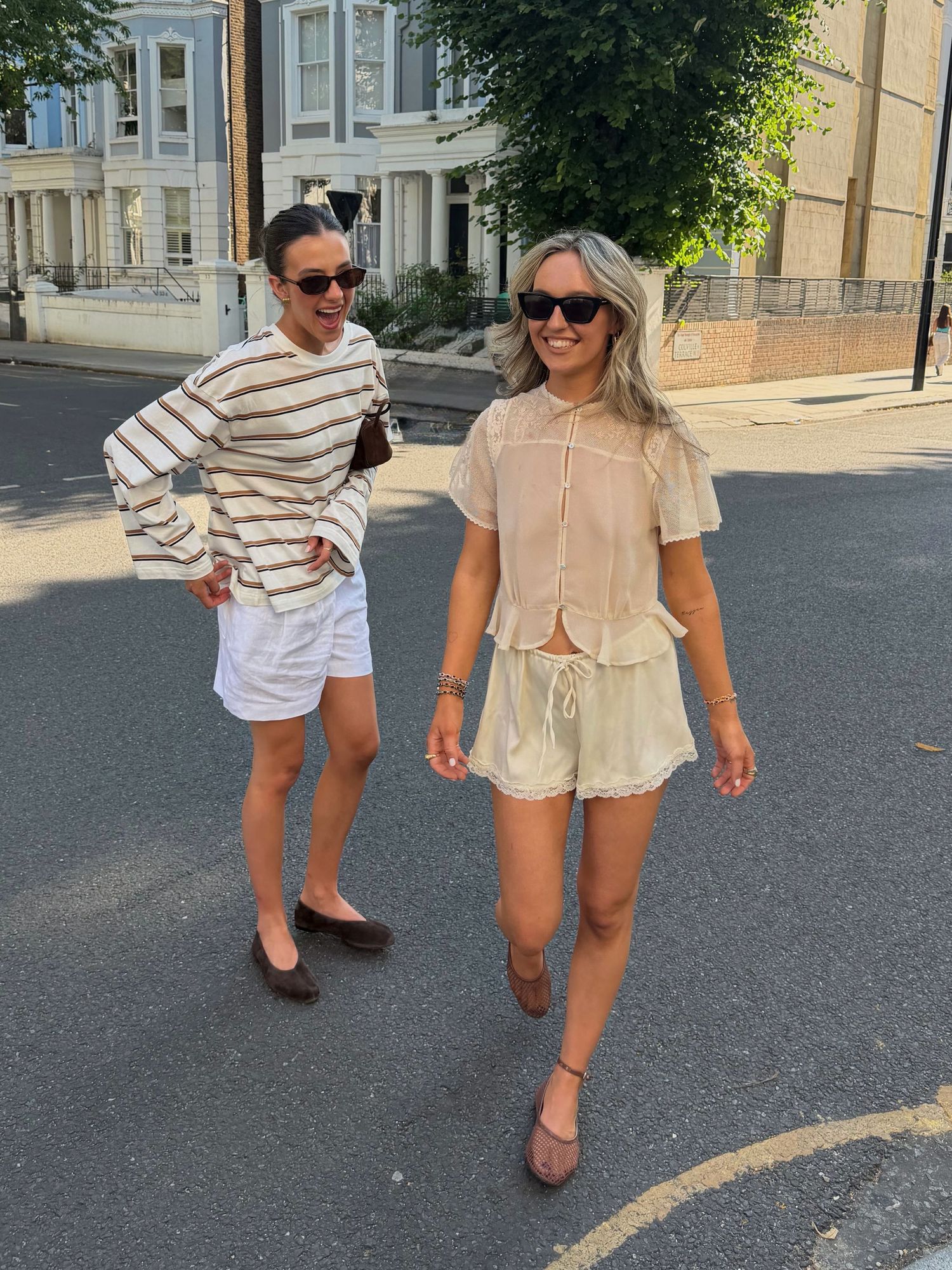 Two women wearing summer outfits walk on a sunny street, one in striped top with white shorts, other in cream blouse and shorts.