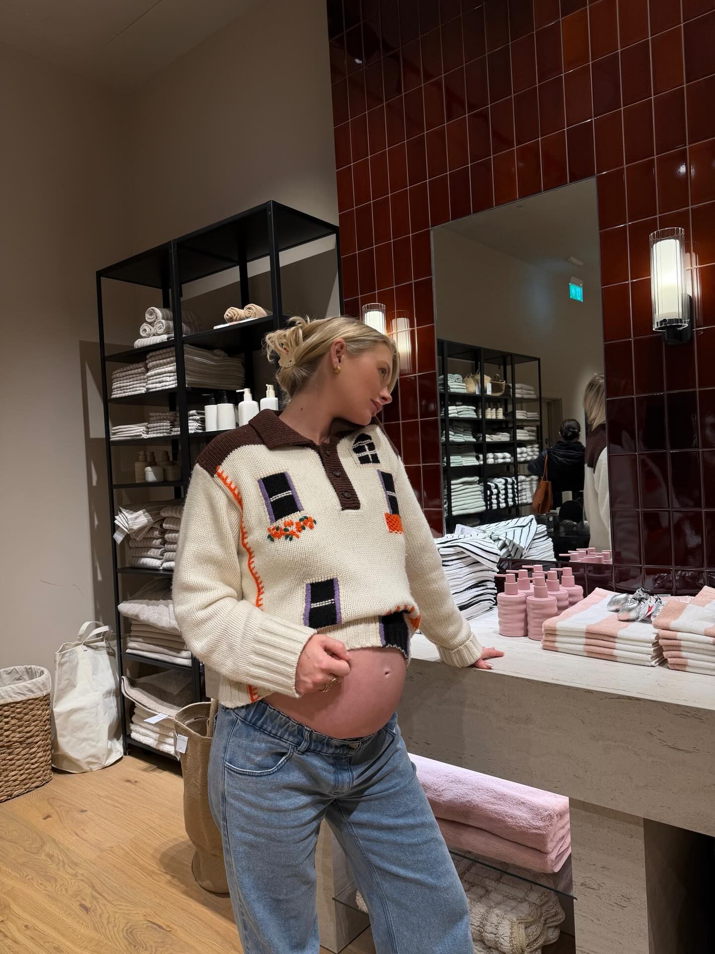 Pregnant woman in cream sweater and jeans touching baby bump in a stylish store with shelves and mirror.