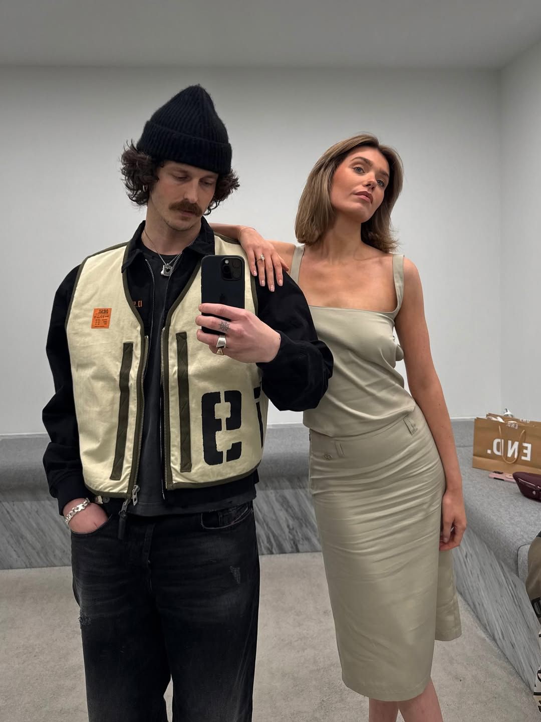 Couple posing for mirror selfie, man in beanie and varsity jacket, woman in elegant sage slip dress.