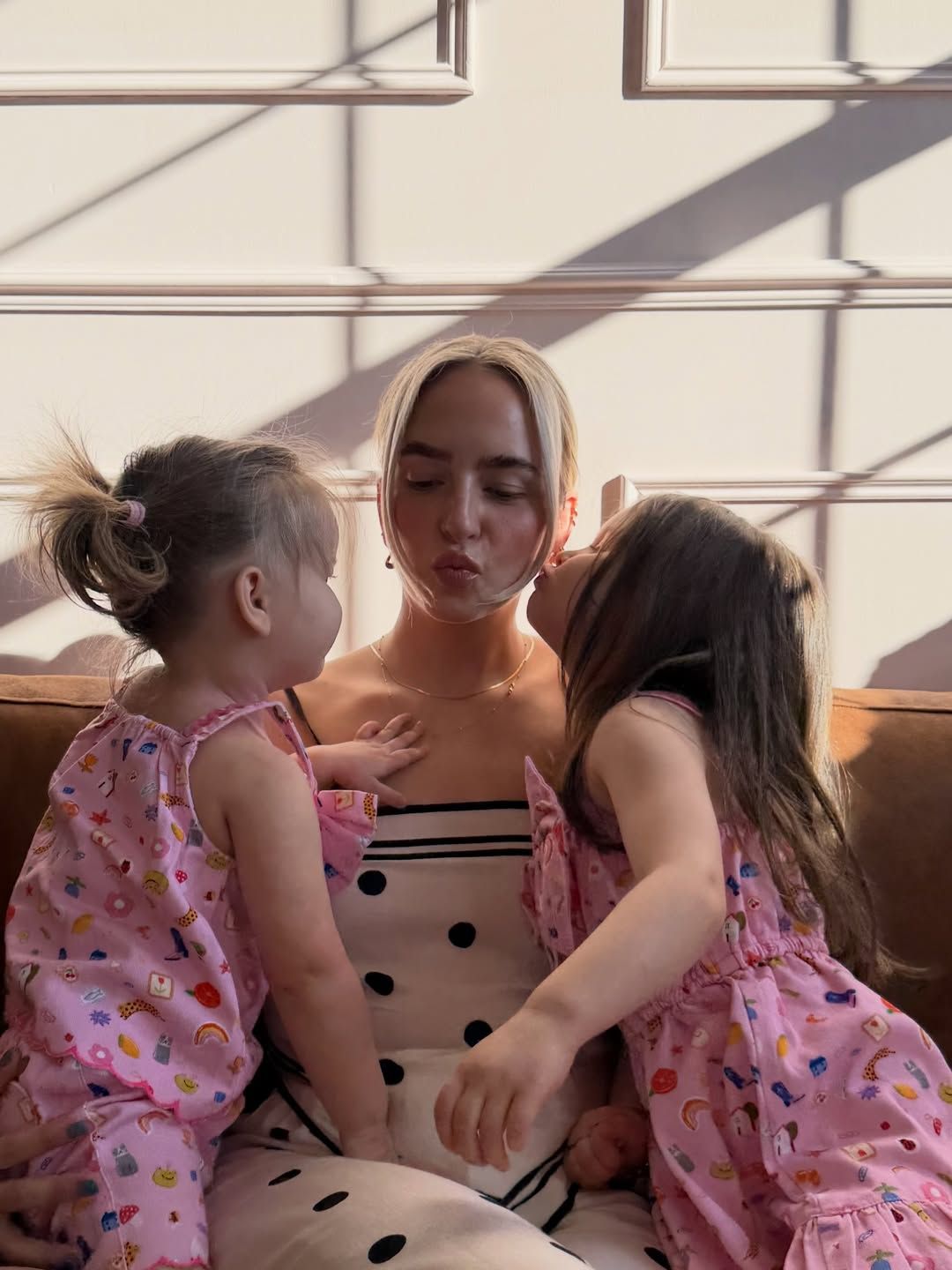 A blonde woman in a polka dot dress sits on a sofa as two young girls in pink floral outfits give her kisses.