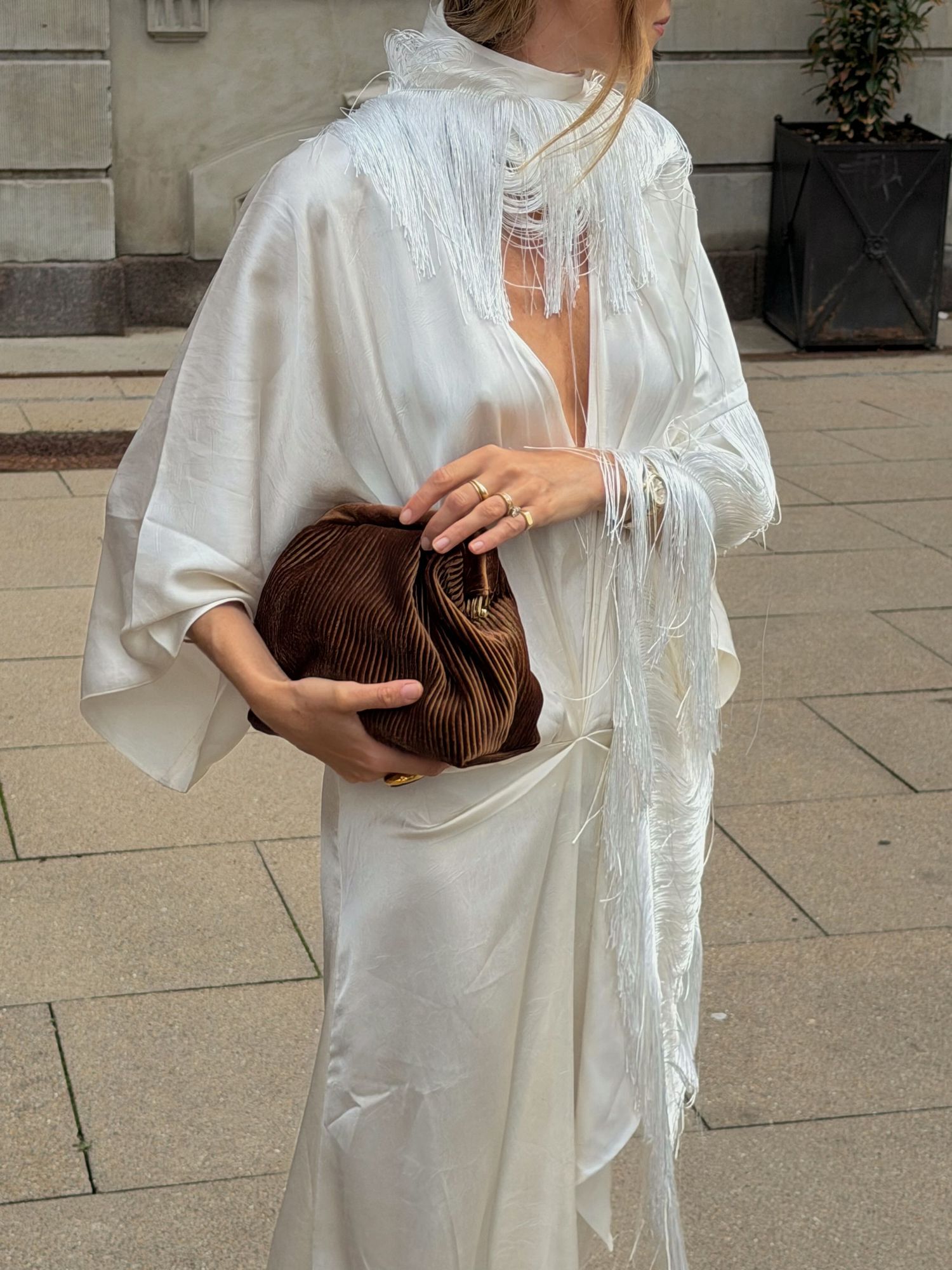 Woman in flowing white satin kaftan with fringe details holding a brown pleated clutch bag outdoors.
