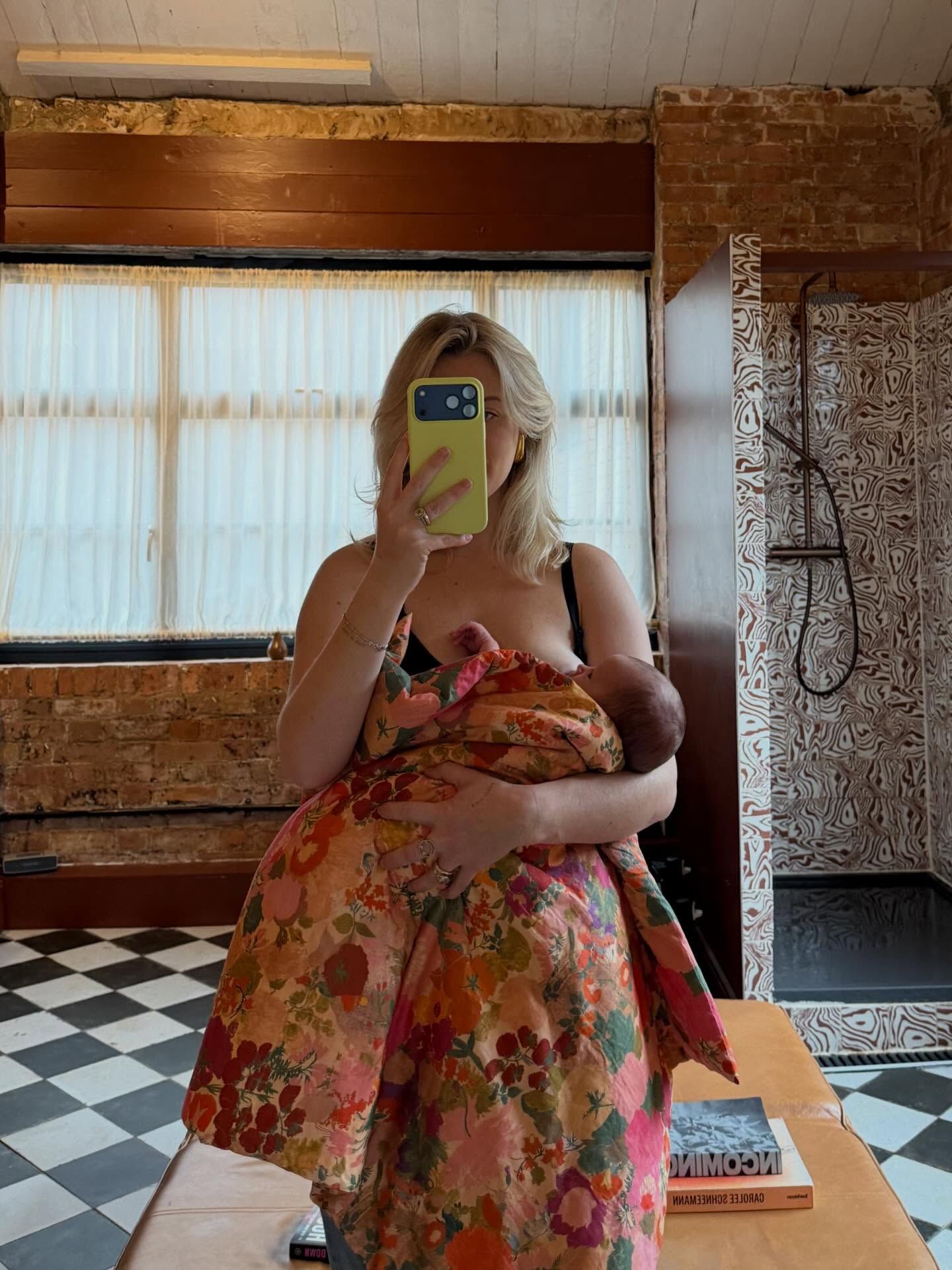 Woman in floral dress holds newborn baby while taking mirror selfie with yellow phone case in stylish bathroom.