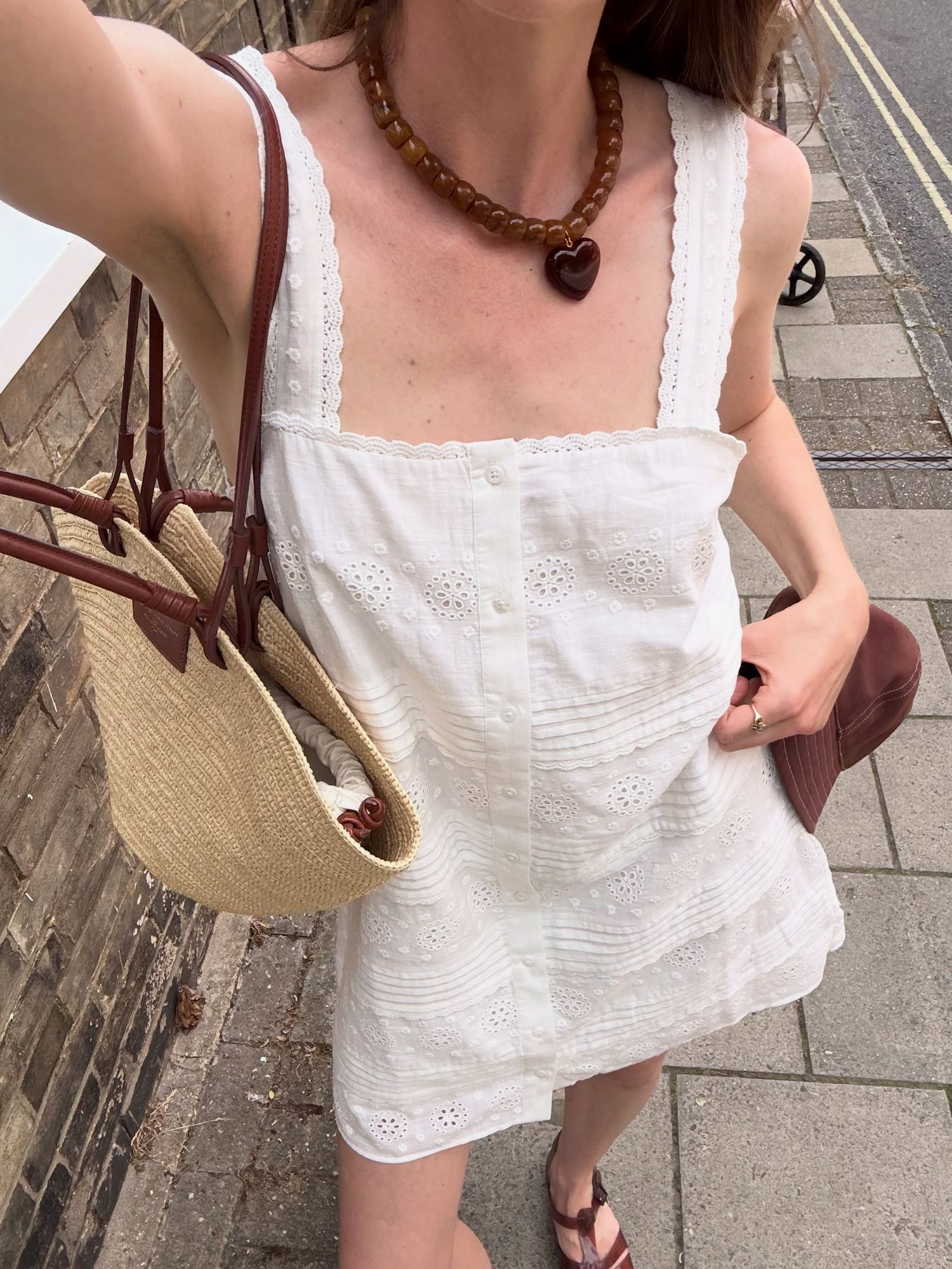 Woman in white eyelet sundress with amber bead necklace, carrying straw tote and suede bag on a sunny sidewalk.