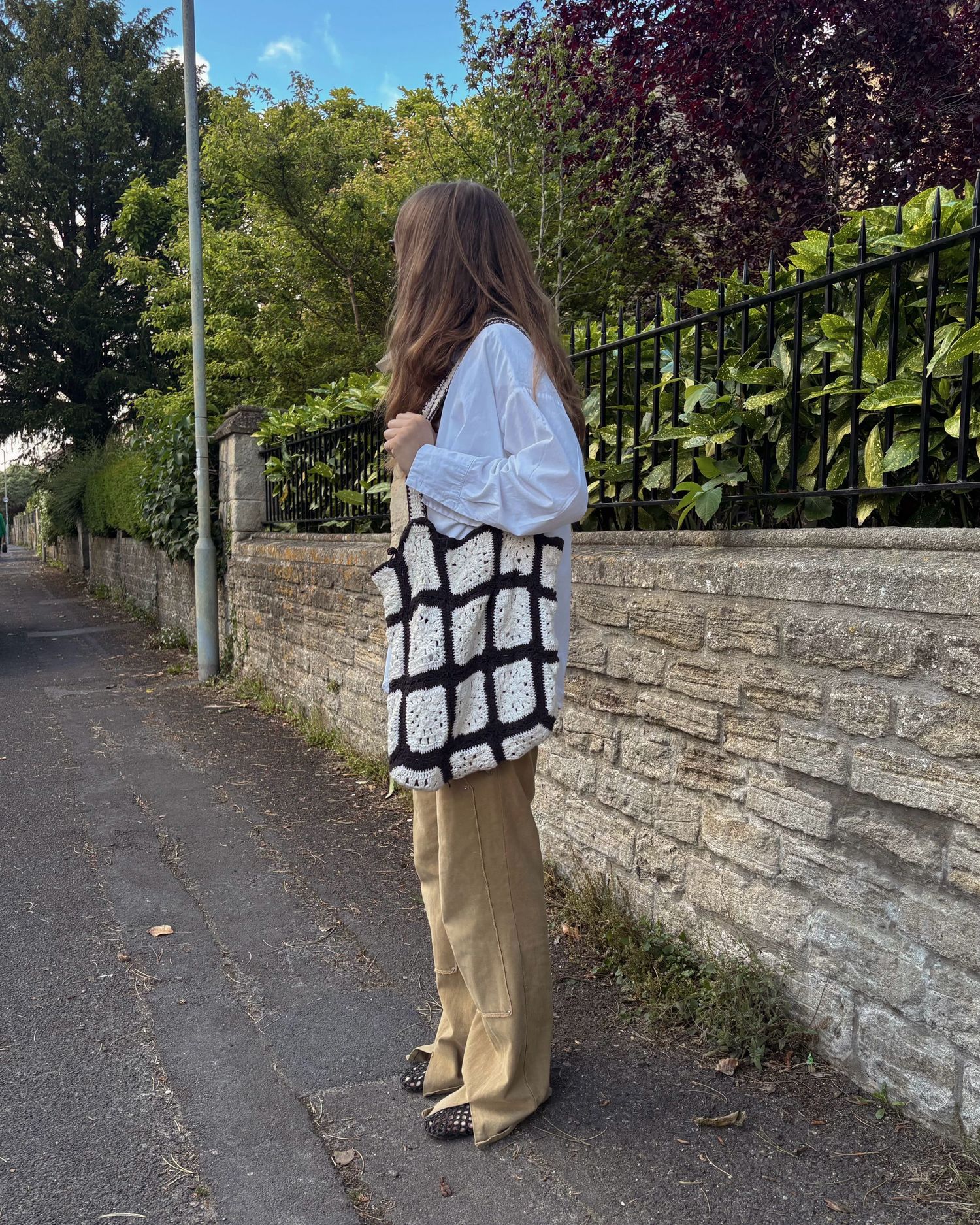 Woman in white blouse, khaki trousers, holding black and white crochet tote bag, standing on a tree-lined sidewalk.