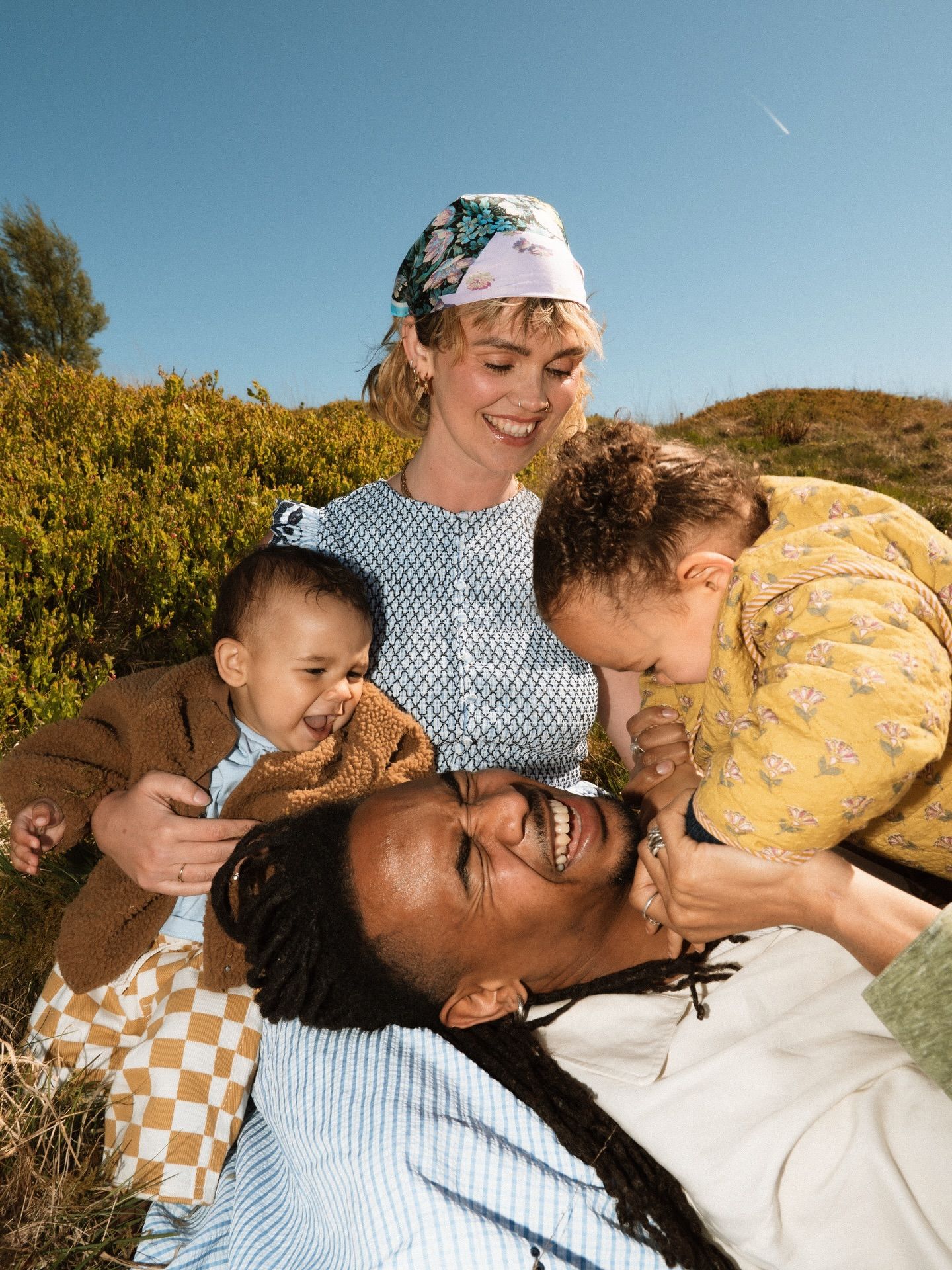 Happy family with two babies laughing outdoors on a sunny day, surrounded by green bushes and blue sky.