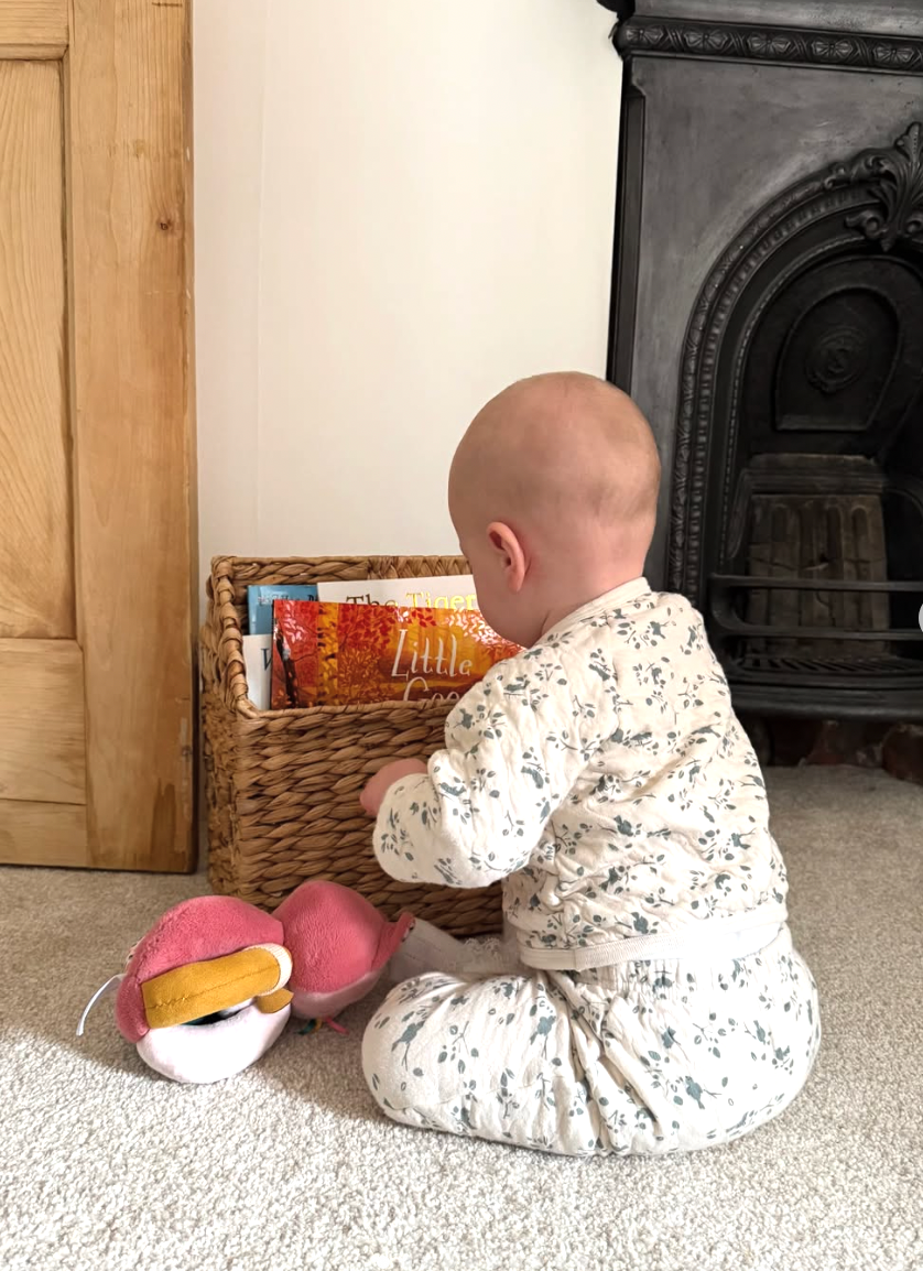 Baby in white polka dot onesie crawls on beige carpet while reaching for toys near a wooden cabinet.