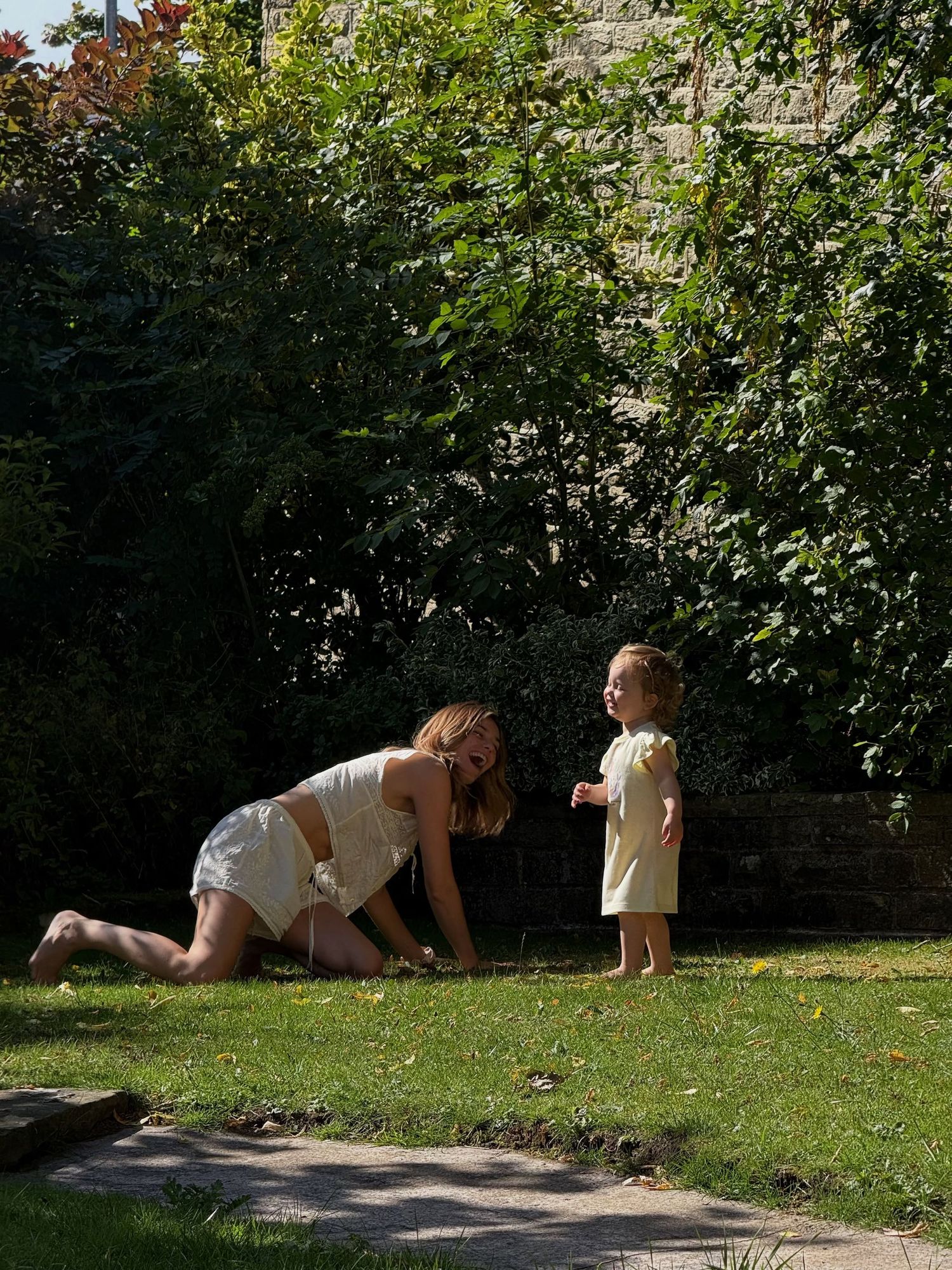 A woman kneeling on grass interacting with a toddler in a sunny garden surrounded by lush green trees.