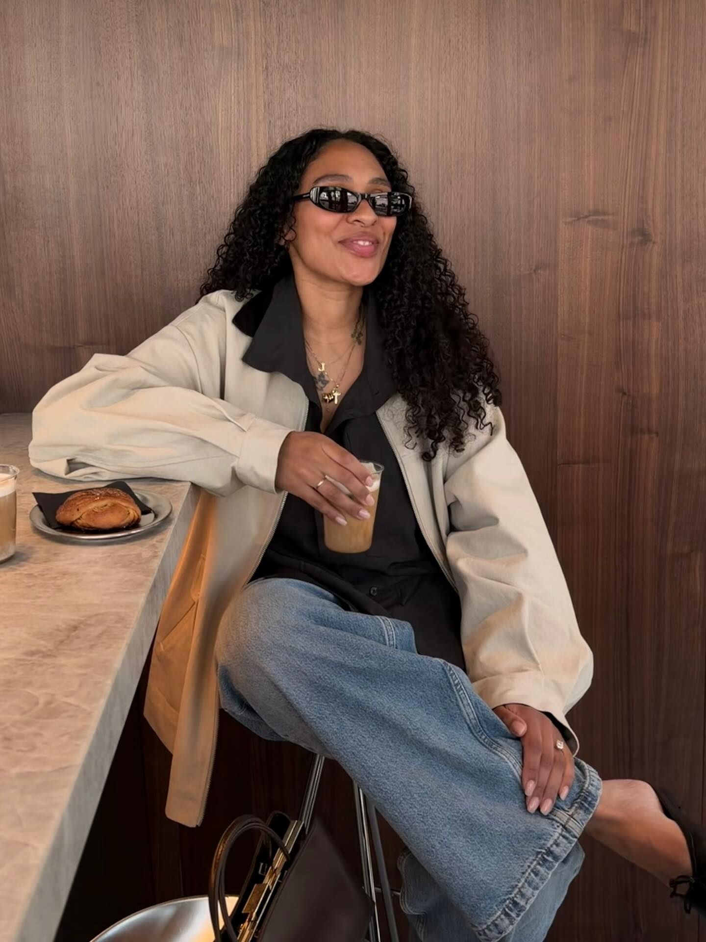 Woman in cream jacket, black top, jeans, and sunglasses sitting at a marble counter with coffee and pastry.