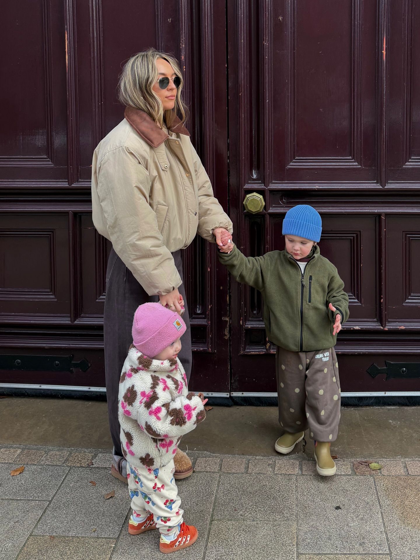 Woman in beige puffer jacket holds hands with toddler boy in blue hat and green jacket, with baby girl in floral outfit nearby.