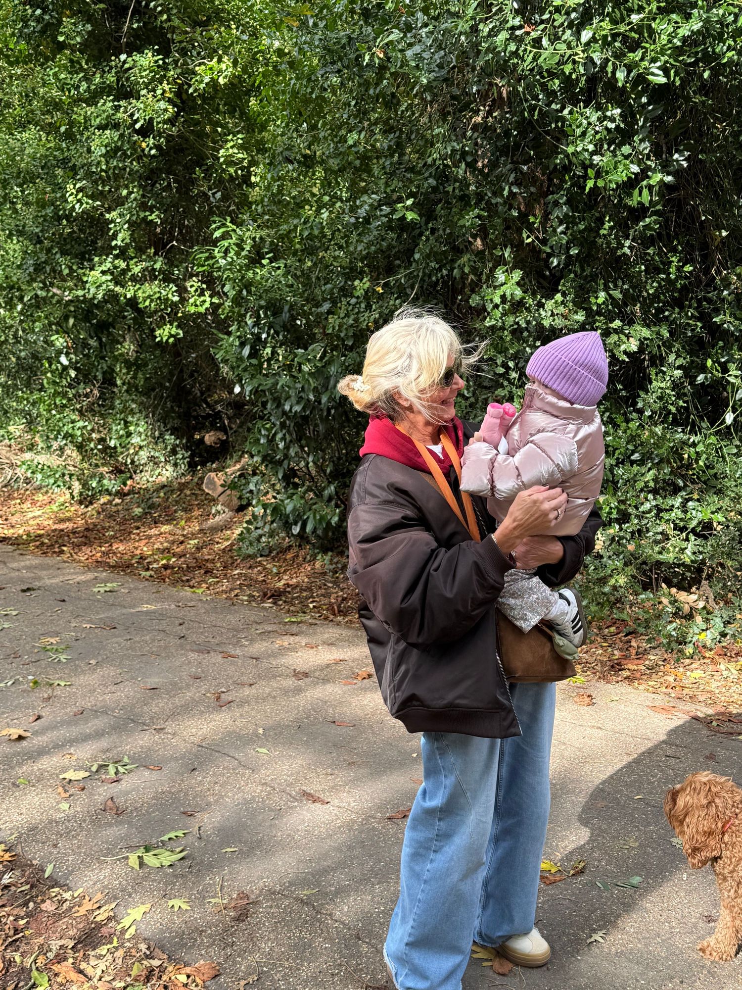 Elderly woman holding a baby in a purple hat on a tree-lined path in autumn.
