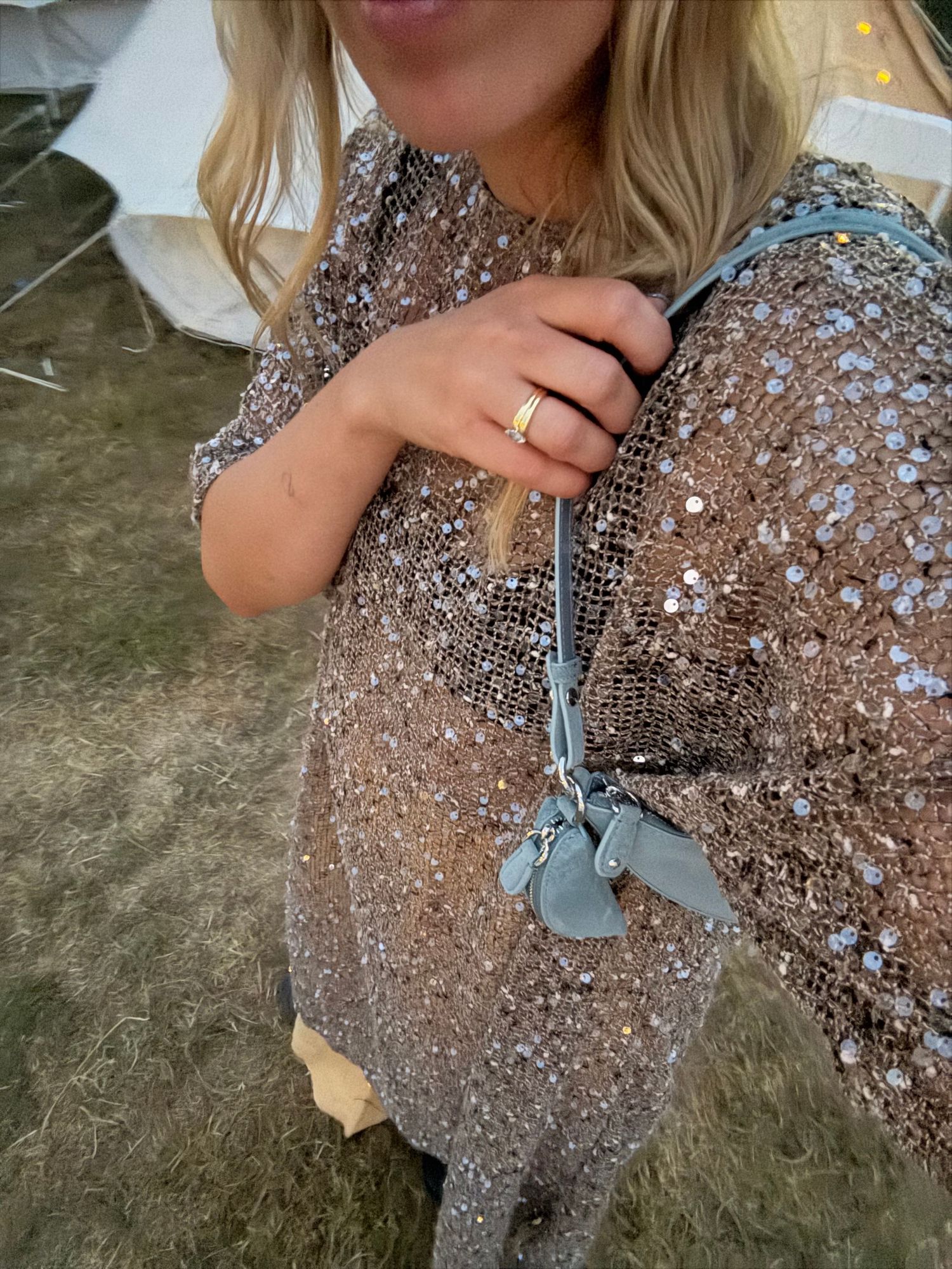 Woman wearing a sparkling rose gold sequin top with a blue crossbody bag and gold ring at an outdoor event.