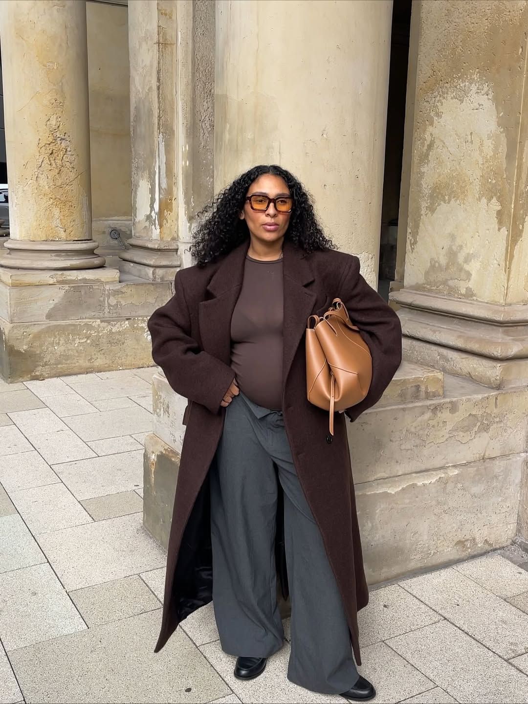 Stylish winter outfit featuring a brown oversized coat, wide-leg trousers, and tan leather bucket bag against stone columns.