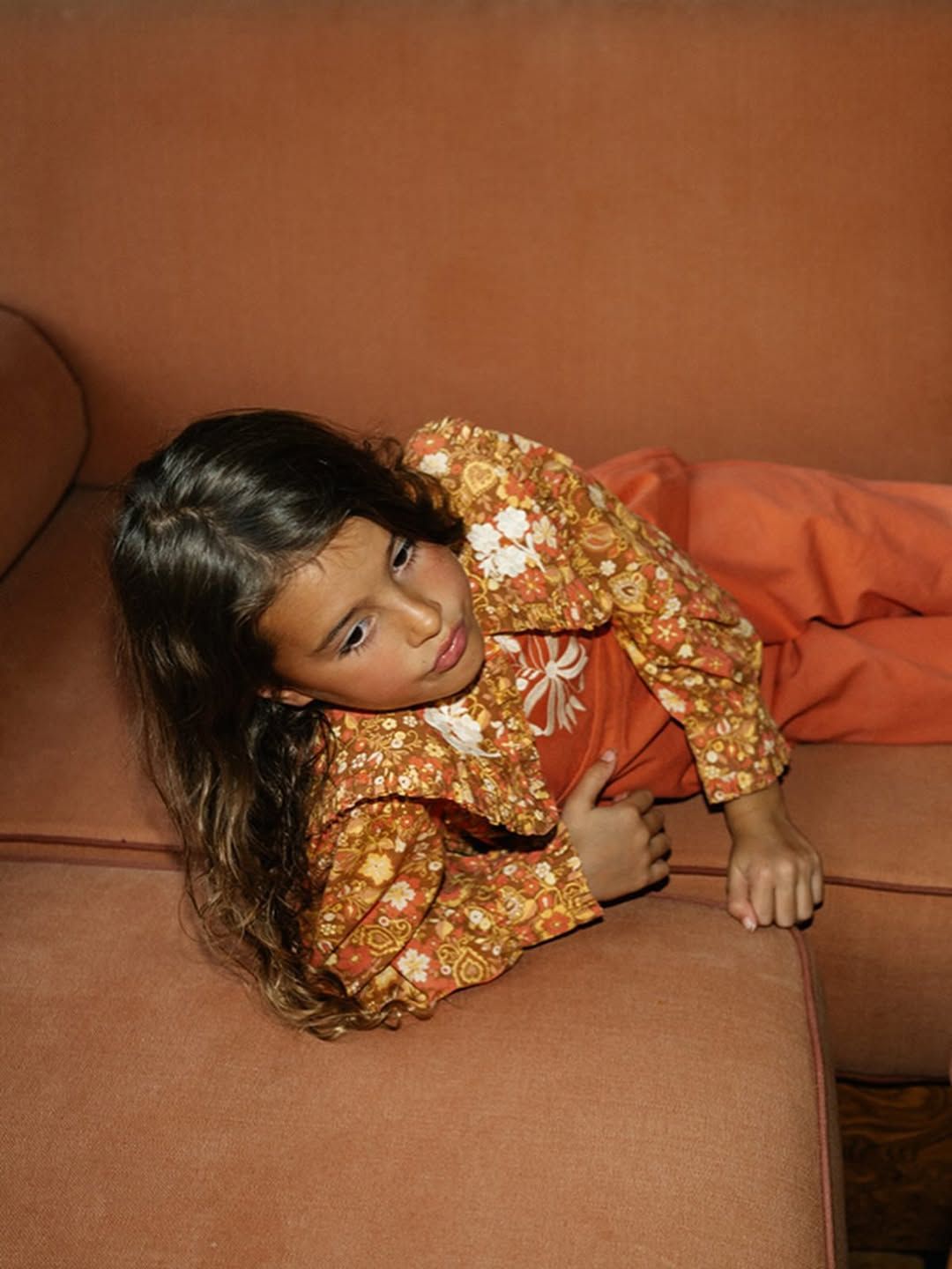 Young girl in floral orange jacket lying on a pink sofa, smiling up at the camera.
