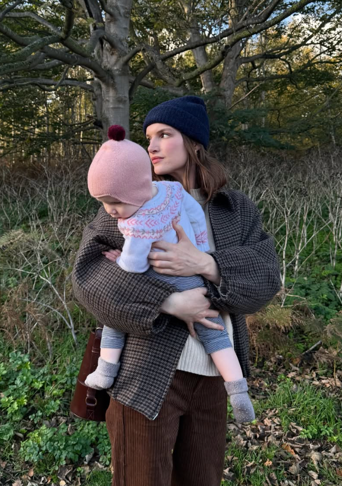 A parent wearing a brown tweed coat holds and comforts a small baby dressed in white on a chilly autumn day outdoors.