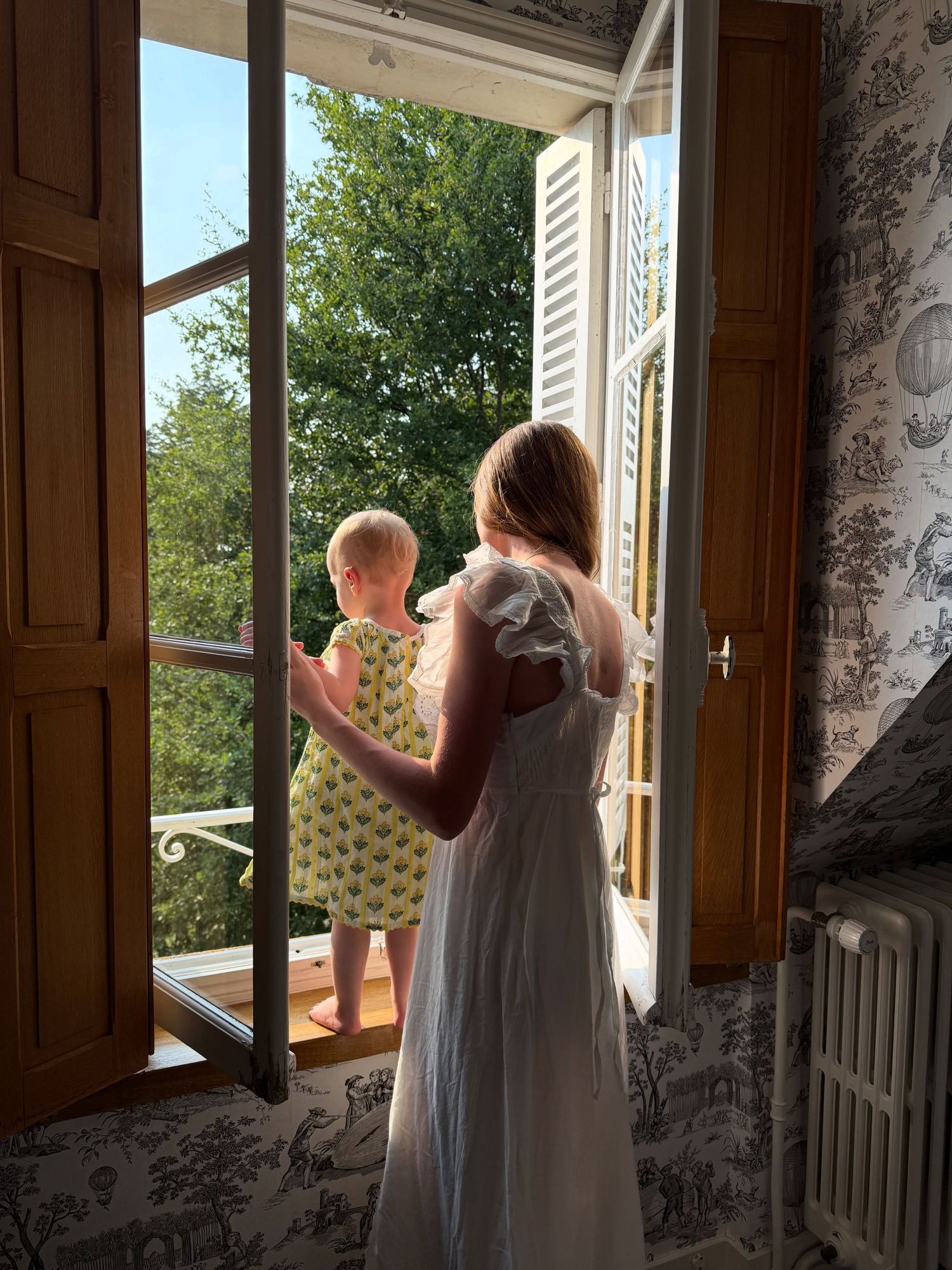 Woman in white dress holds baby near open French doors overlooking lush garden in elegant room with toile wallpaper.