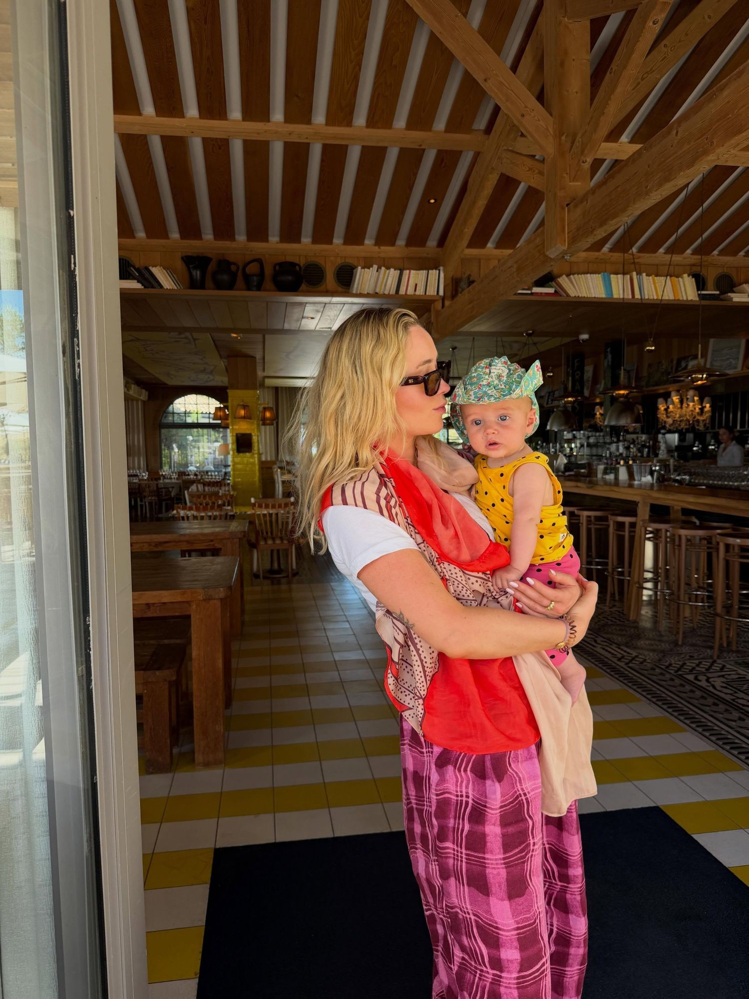 Woman in colorful outfit holding a baby in yellow dress inside a tropical restaurant.
