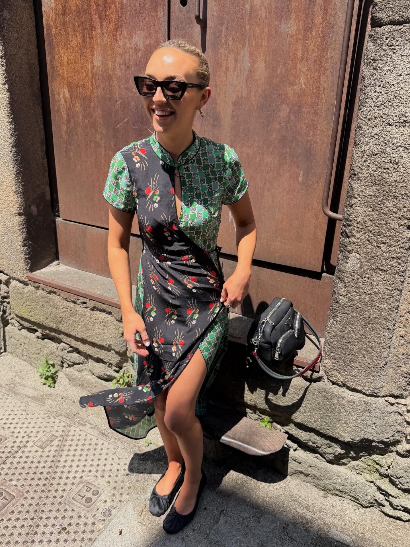 Smiling woman in patterned green dress with sunglasses holding bag on cobblestone street.