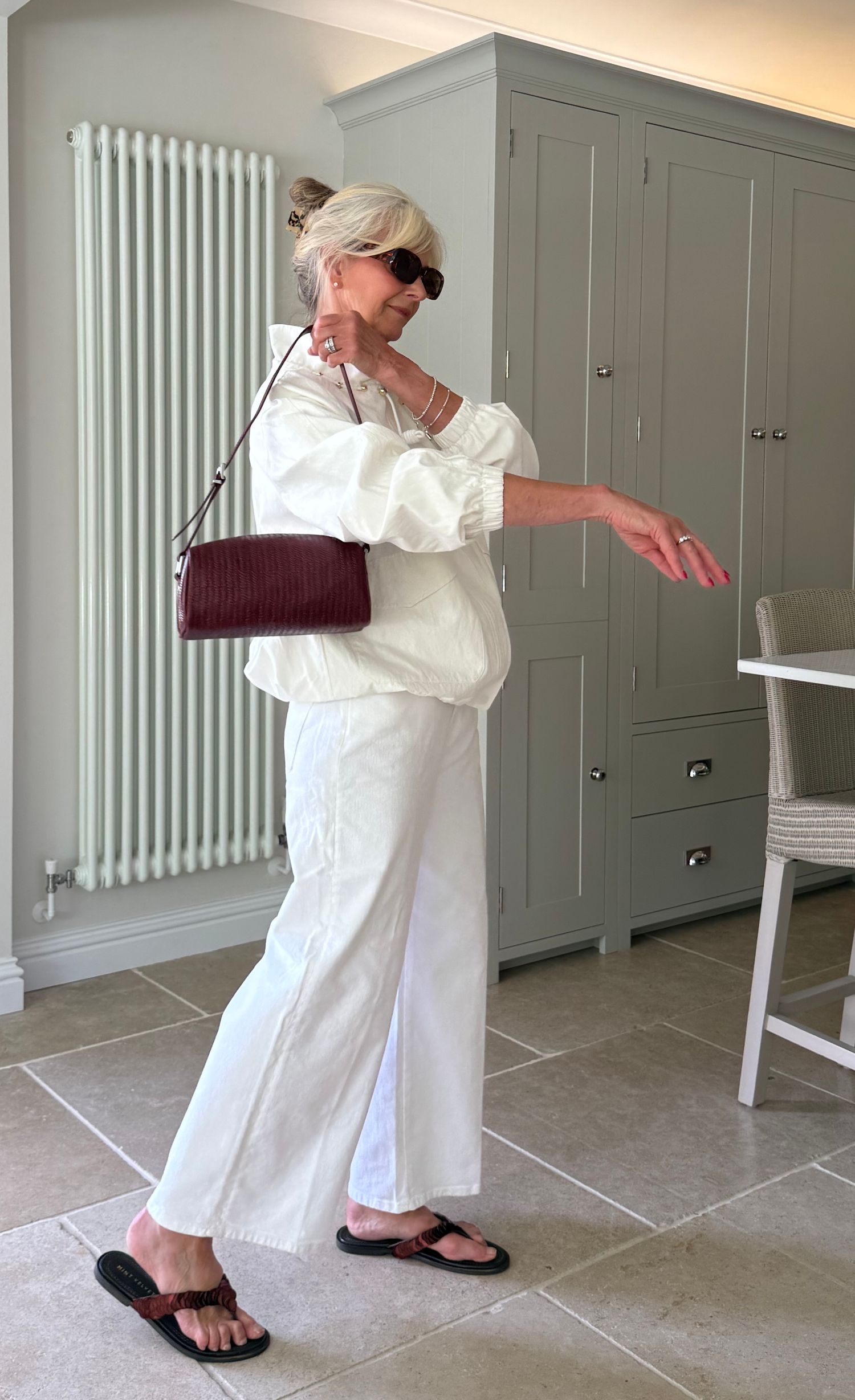 Woman in white linen outfit with burgundy shoulder bag, black sandals, and sunglasses posing indoors.