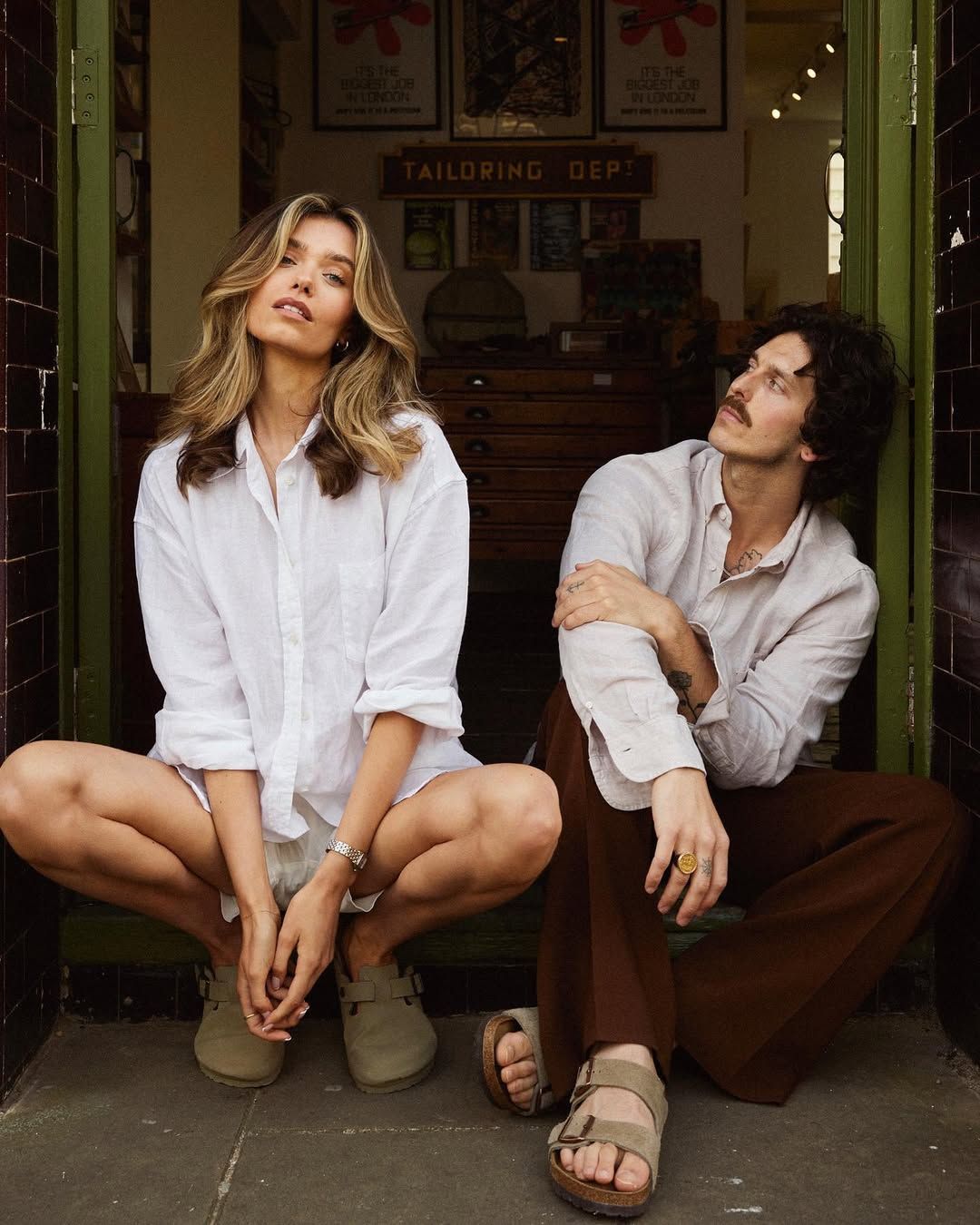 Man and woman in casual linen outfits sitting together outside a tailoring shop doorway.