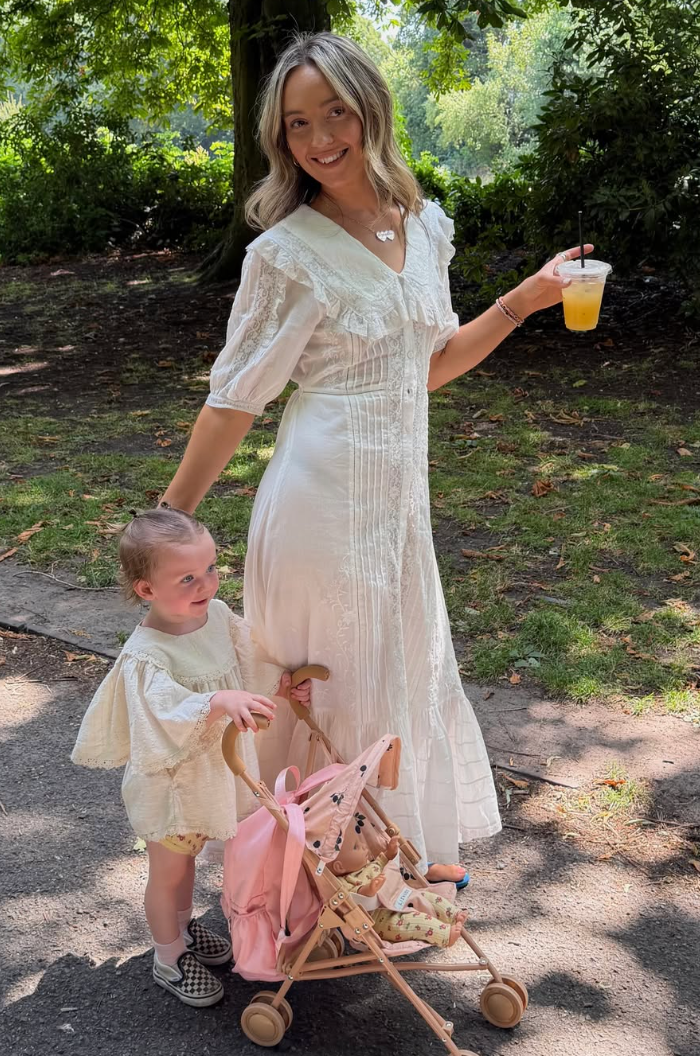 Woman in white lace dress holding drink with toddler girl pushing pink toy stroller in sunny park.