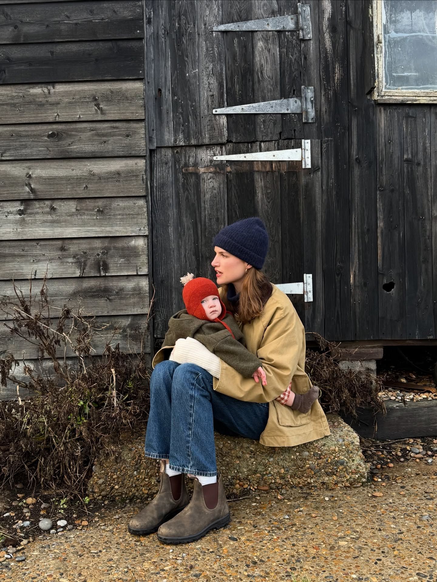 A woman in a tan jacket and navy beanie sits outdoors holding a baby in a red hat near a weathered wooden barn.