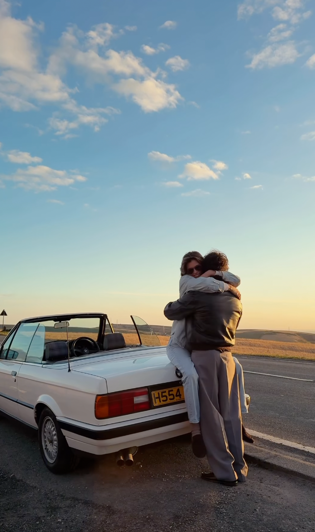 Couple embracing romantically beside a white convertible BMW at sunset on an open road.