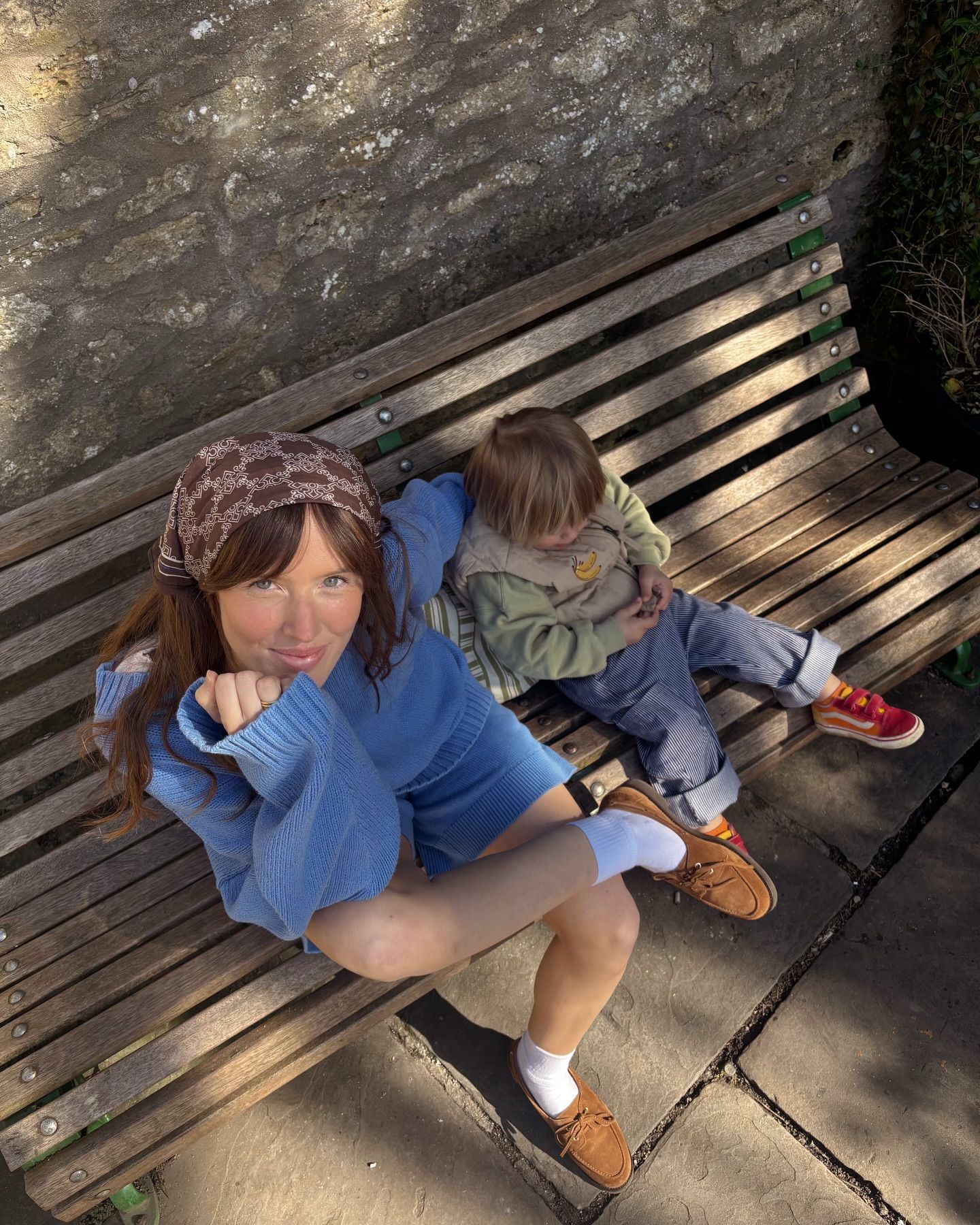 Woman in blue outfit and headband sits on wooden bench with a toddler in a sunny outdoor setting.