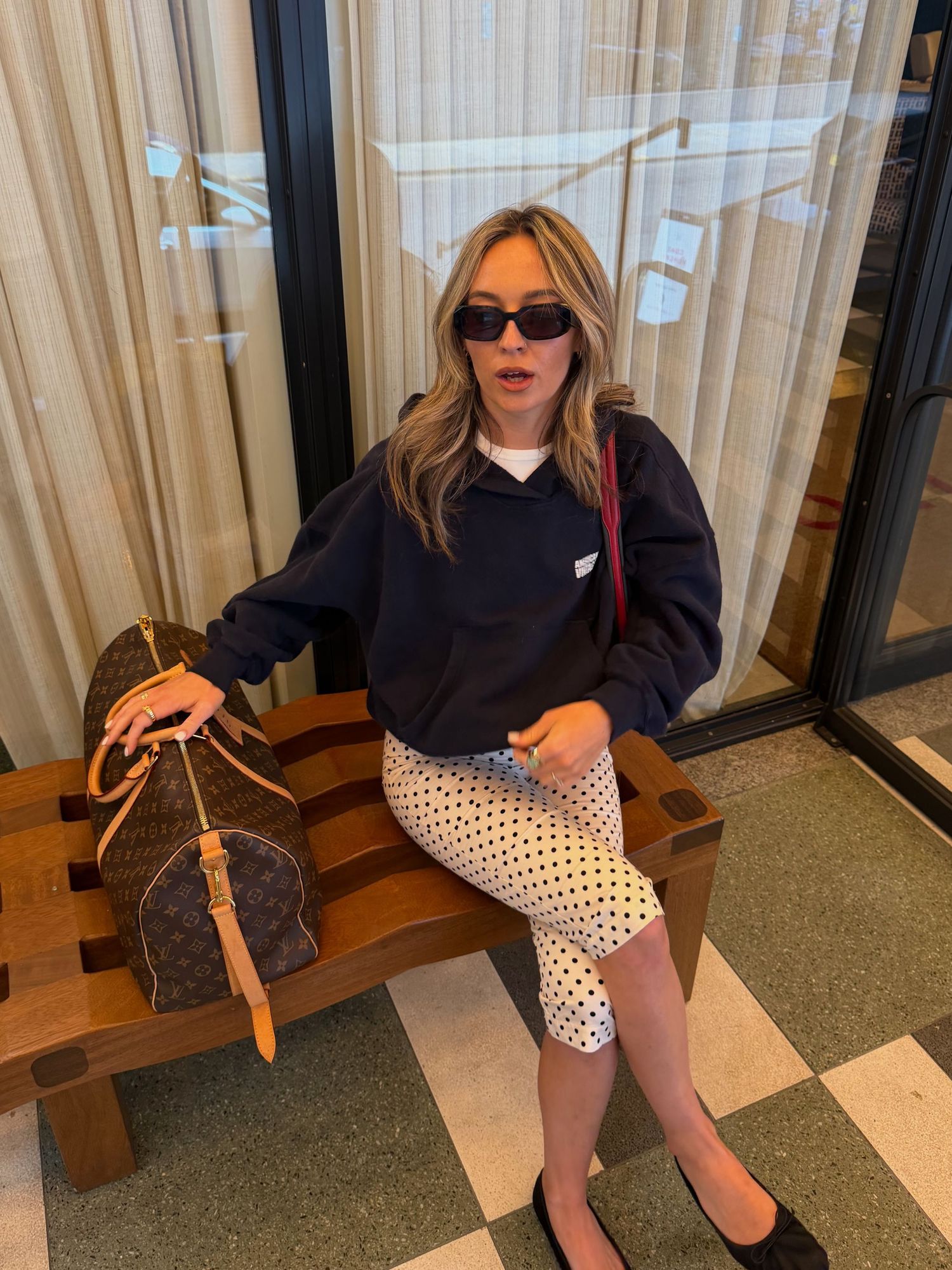 Woman in black hoodie and polka dot skirt sitting on bench with Louis Vuitton bag, wearing dark sunglasses.