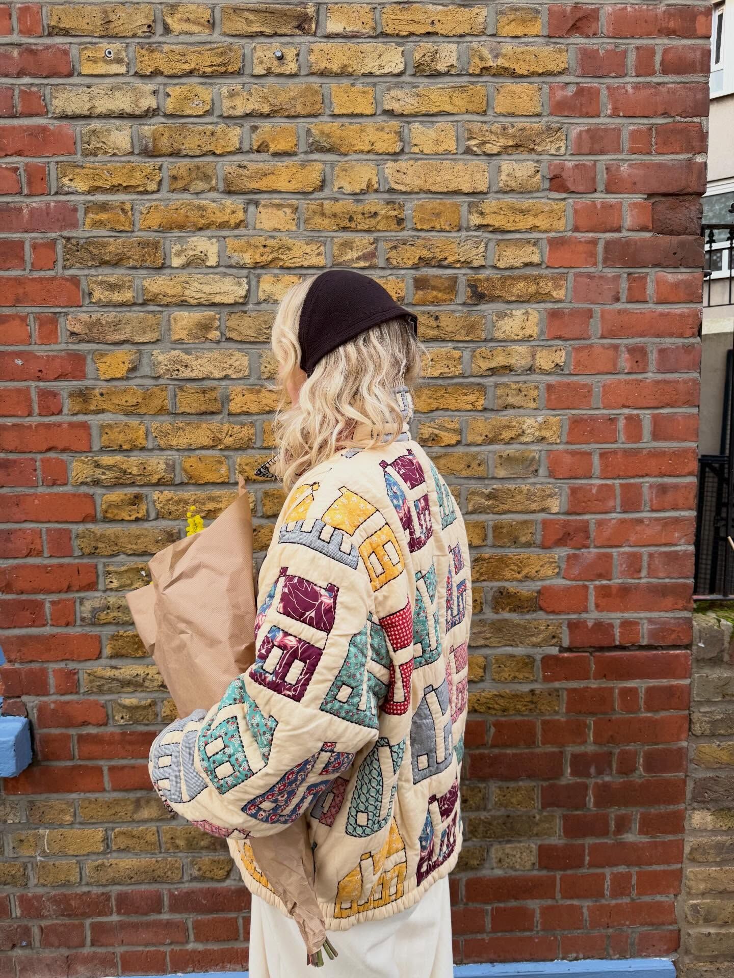 Woman in colorful quilted jacket with house print, black beanie, holding brown bag, standing by brick wall.