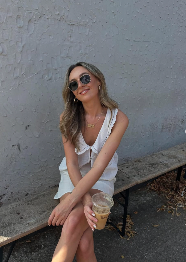 Fashionable woman in white summer dress and sunglasses sits outdoors holding an iced beverage against grey wall.