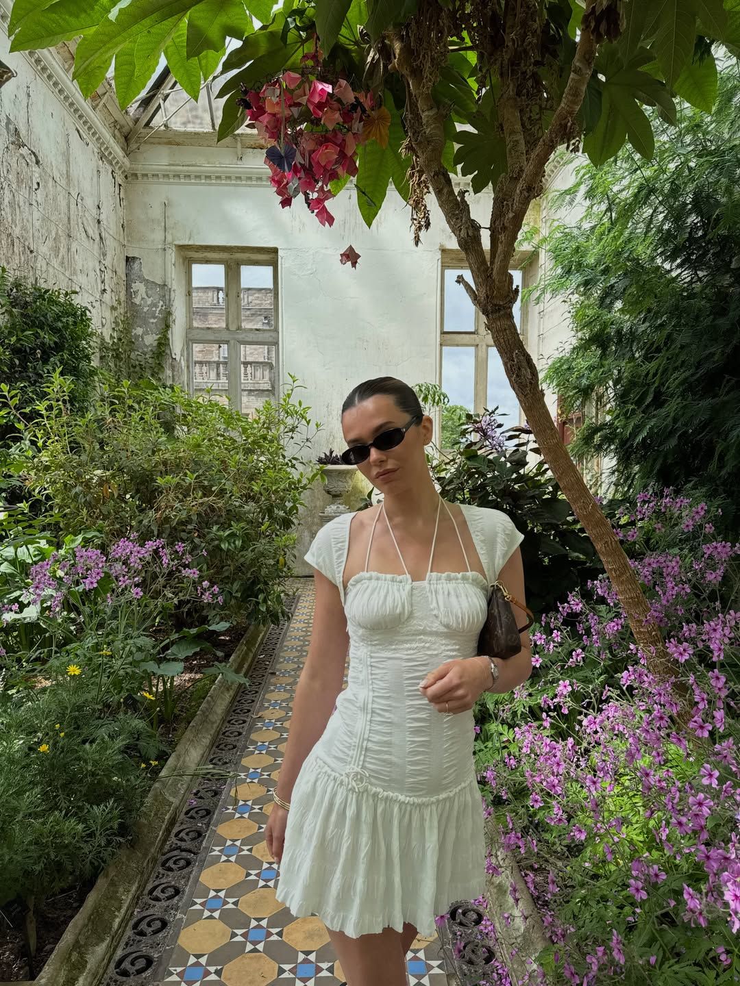Woman in white ruched mini dress and sunglasses posing in a lush garden courtyard with colorful flowers and mosaic tiles.