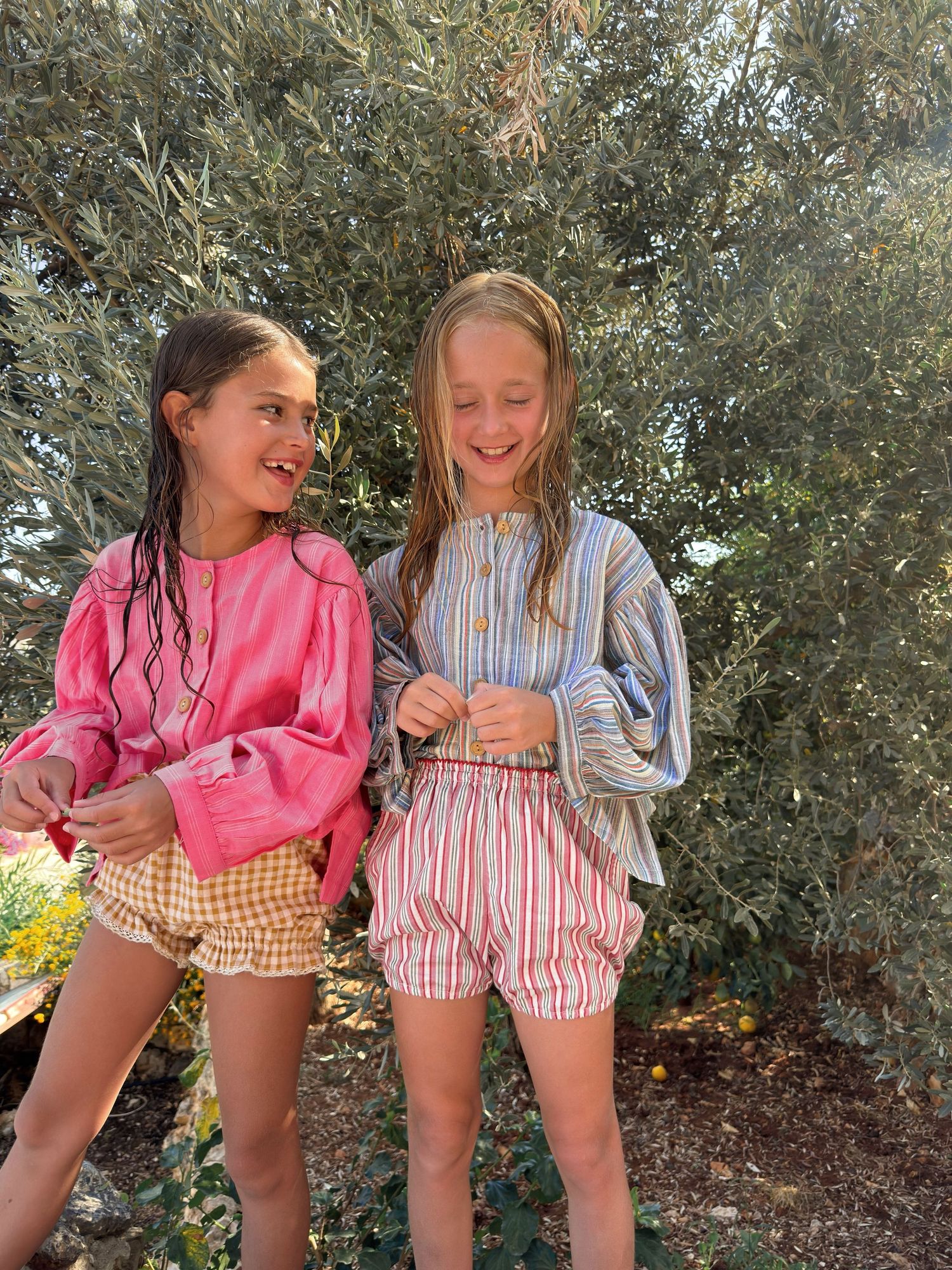 Two young girls with wet hair smile outdoors, wearing colorful summer outfits with striped and gingham shorts.