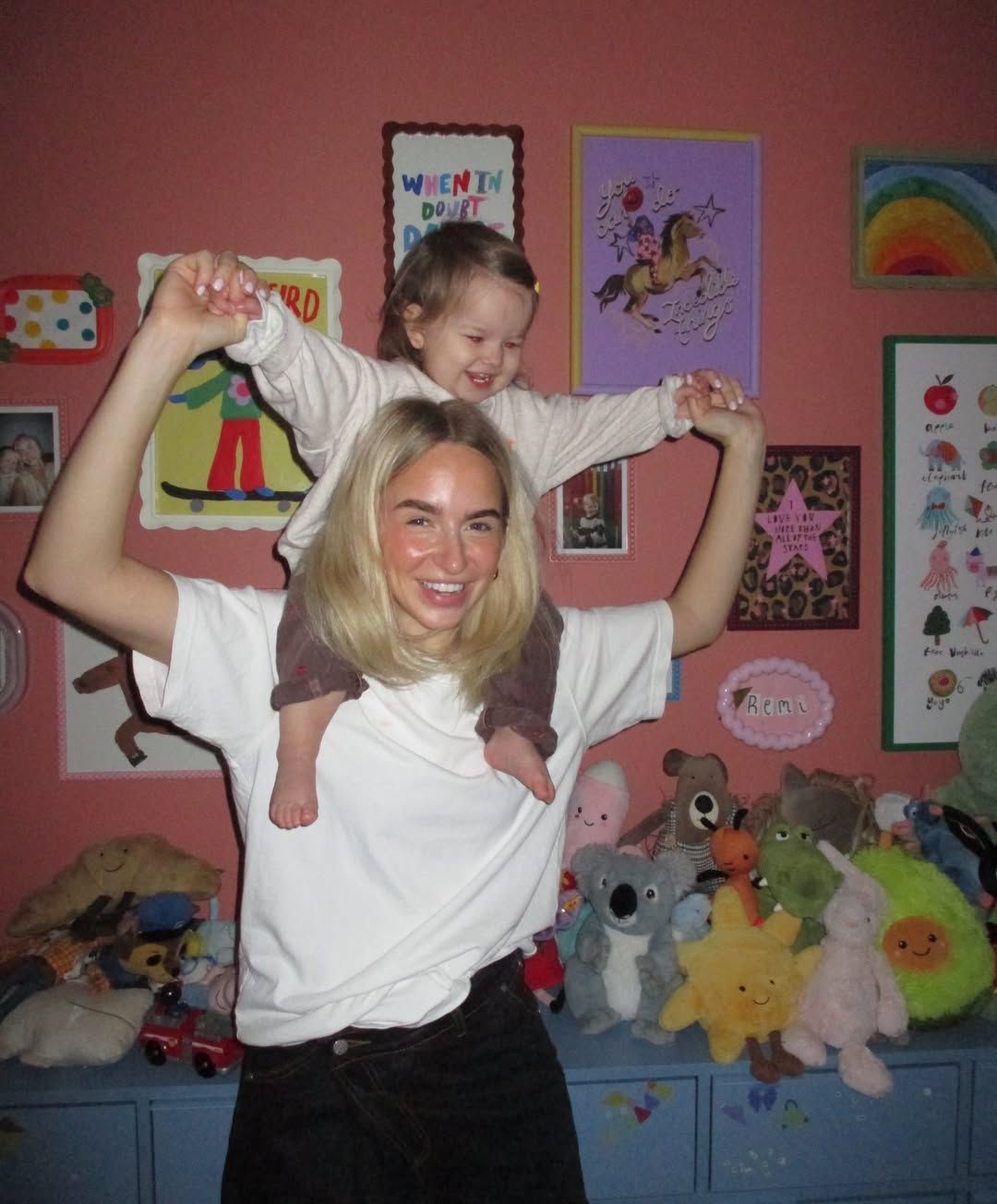 Smiling blonde woman with toddler on her shoulders in colorful kids room with stuffed animals and posters.