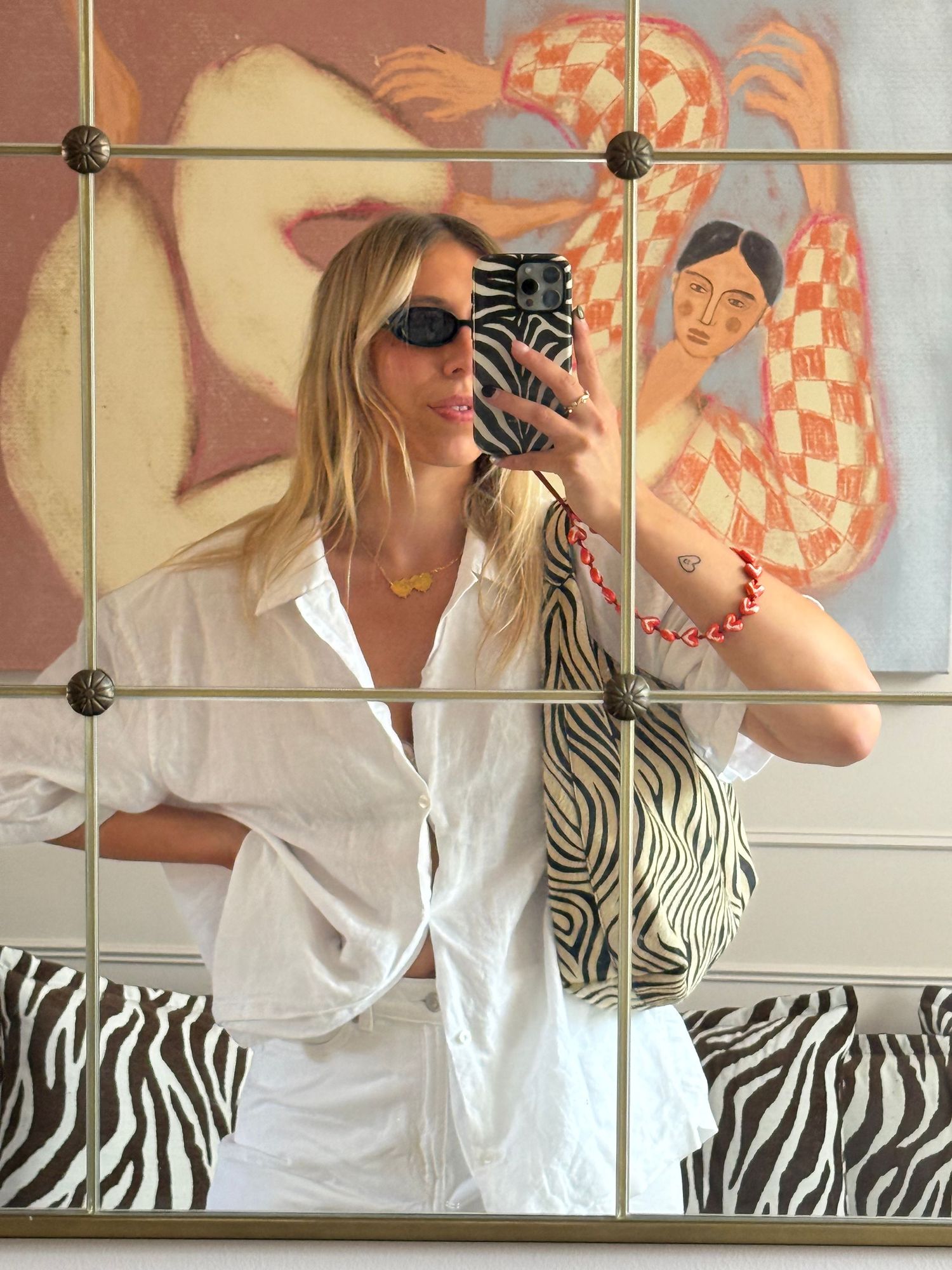 Woman in white oversized shirt takes mirror selfie holding zebra print bag, wearing sunglasses and gold necklace.